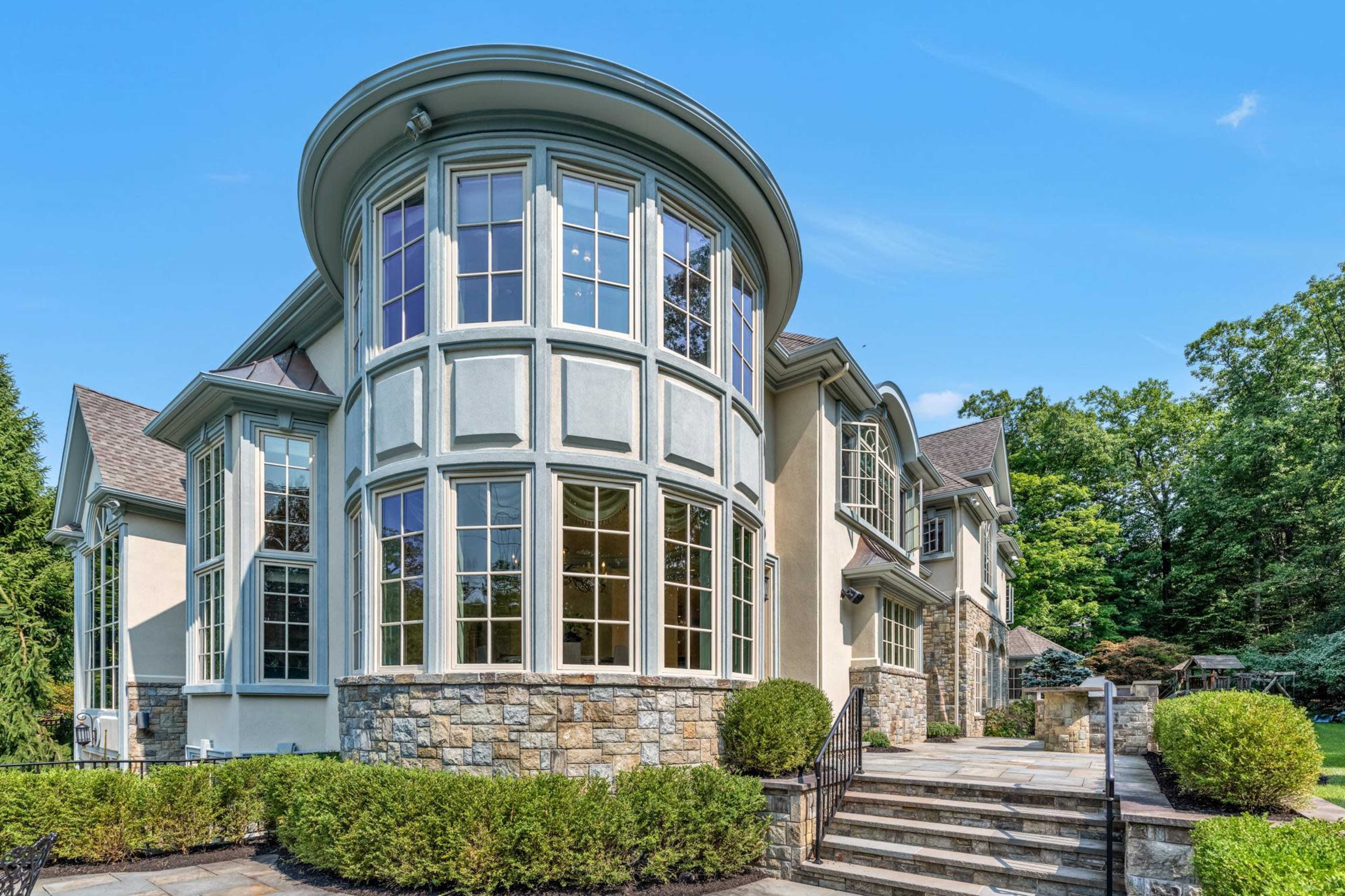 The image shows a large, modern house with a round, bay window and a stone facade surrounded by manicured hedges and a paved walkway.