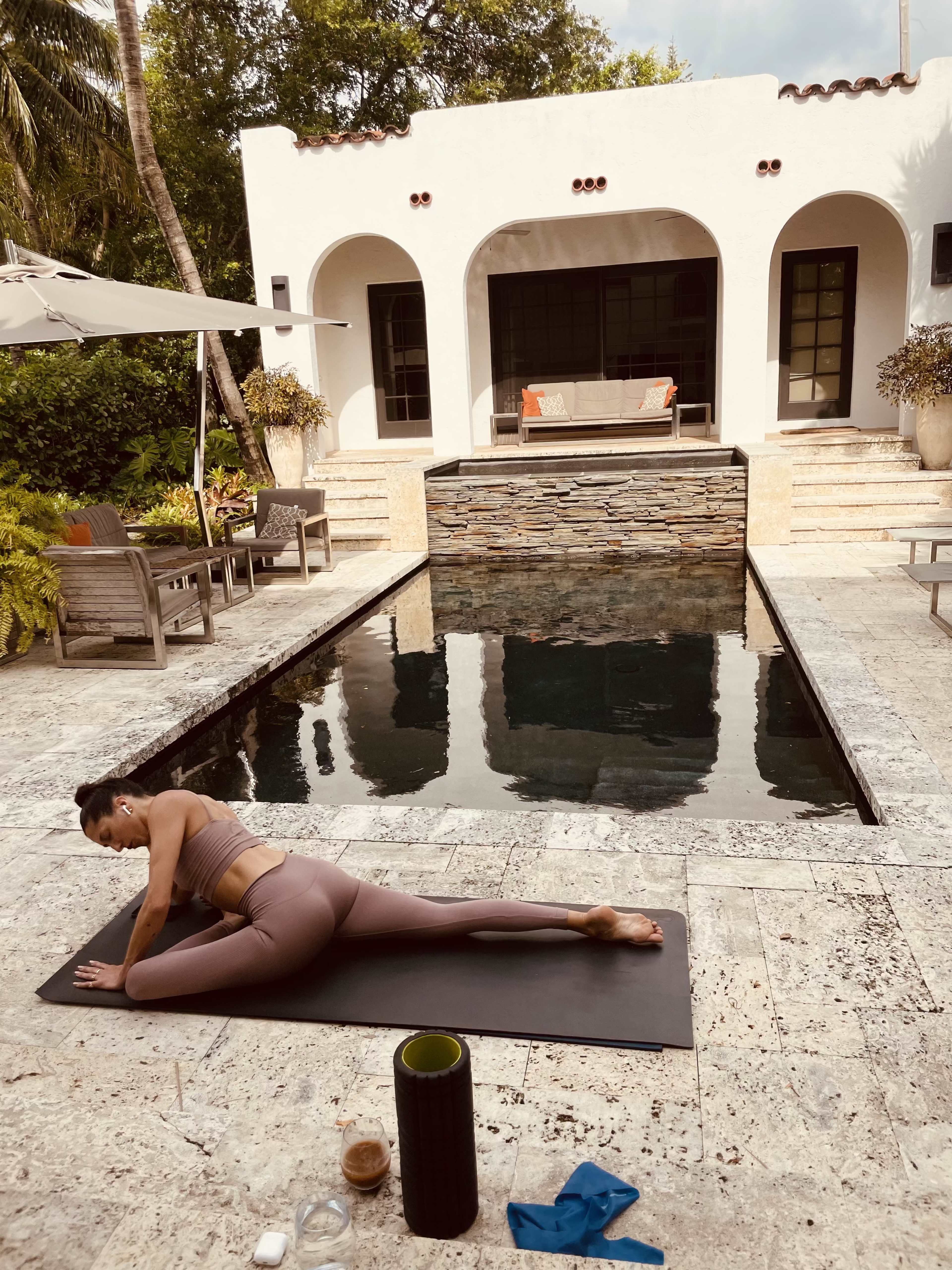 A person performs a yoga pose on a mat beside a pool in a serene outdoor setting featuring a modern home with arched windows and lush greenery.