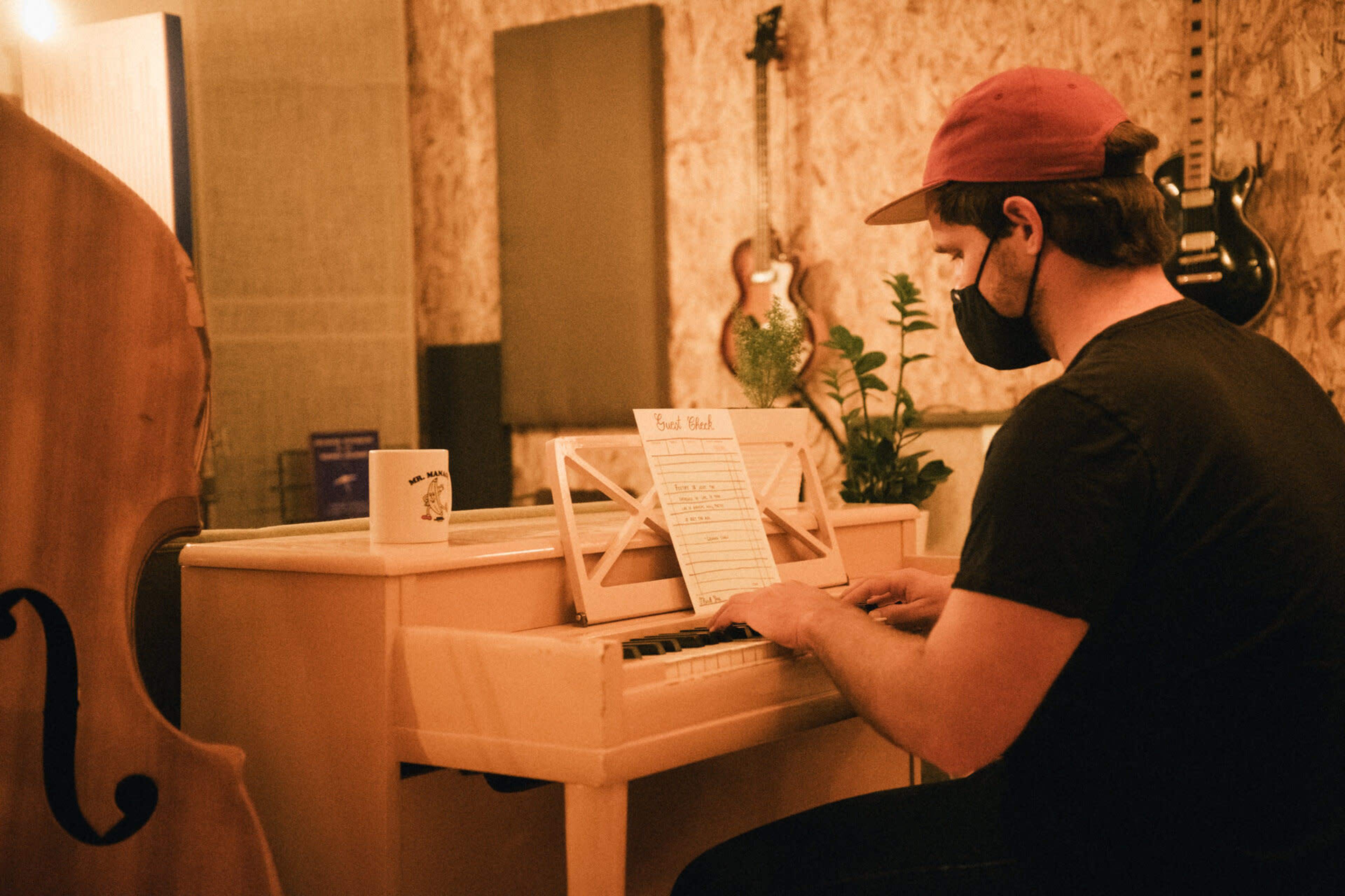 A person wearing a mask and a red cap plays a light-colored piano in a music studio surrounded by instruments.