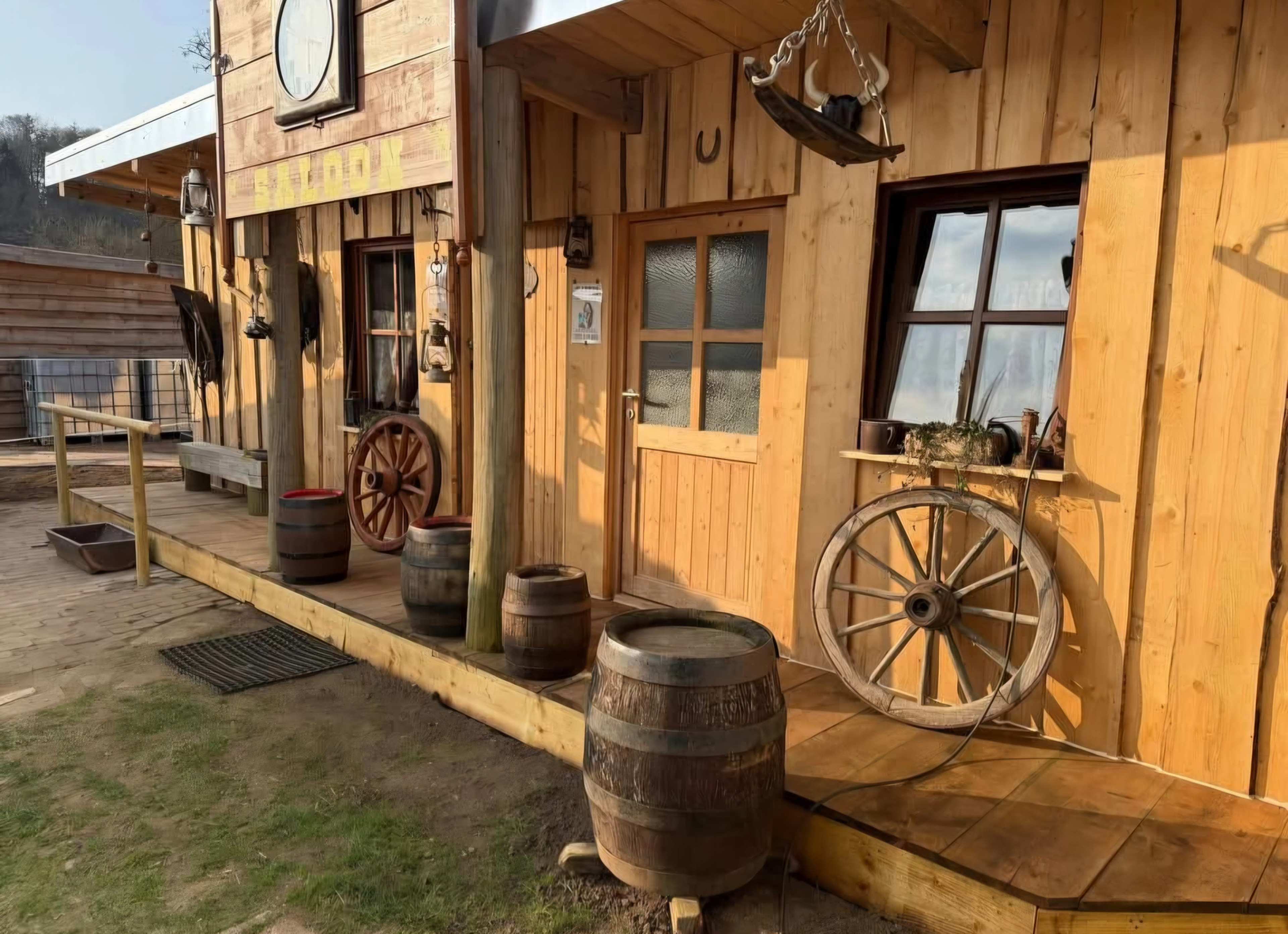 The image shows a rustic wooden building with a porch, barrels, a wagon wheel, and a clock mounted on the facade.