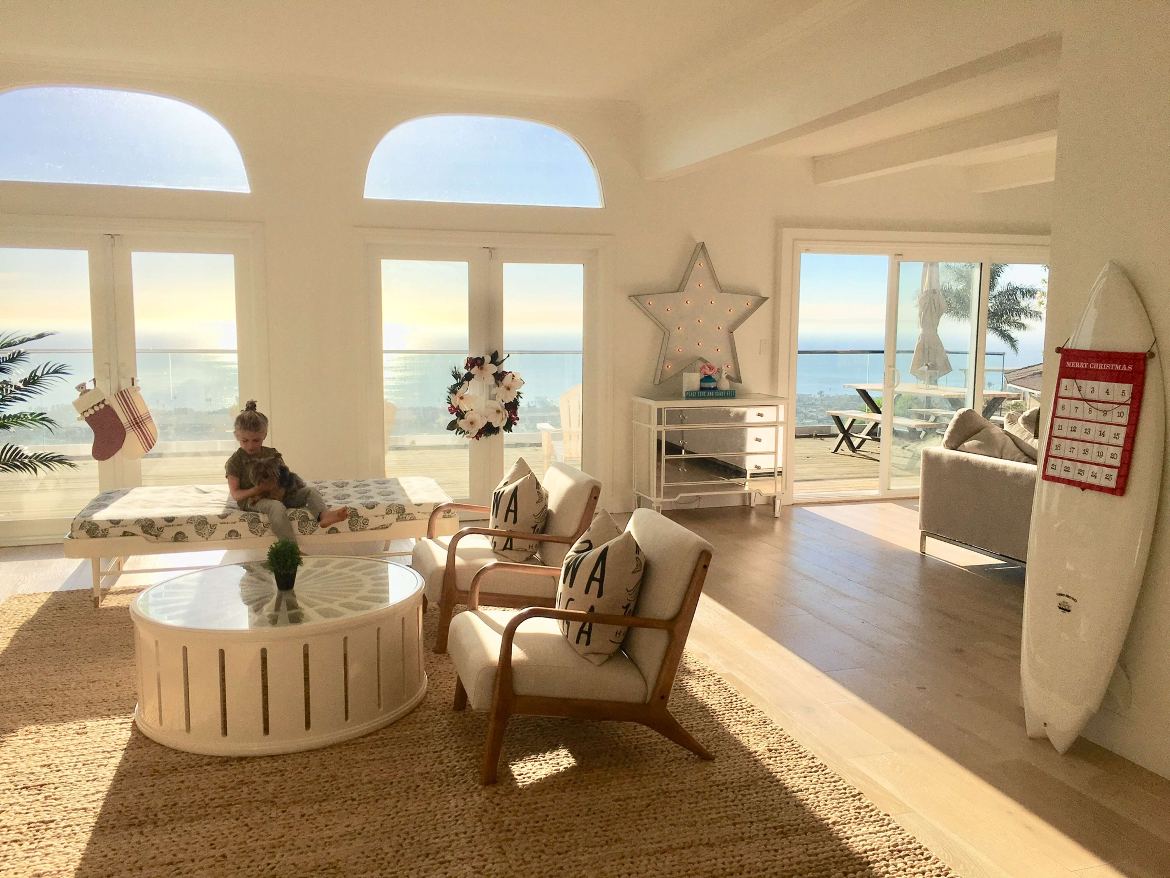 A bright, airy living room features a child sitting on a bench beside a circular coffee table, with large windows showcasing a view of the ocean.