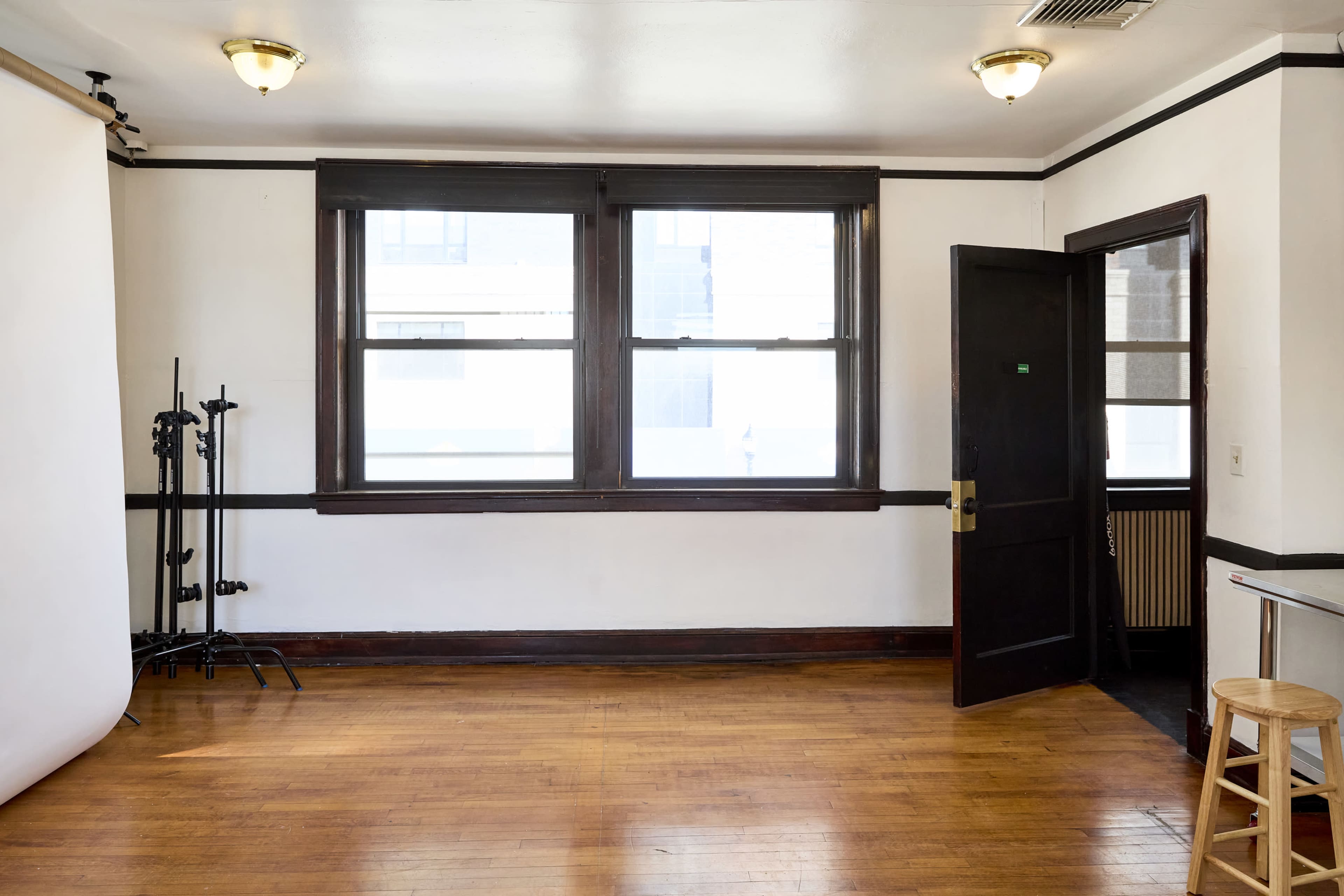 The image shows a sparsely furnished room with wooden flooring, two large windows, and a door leading outside.