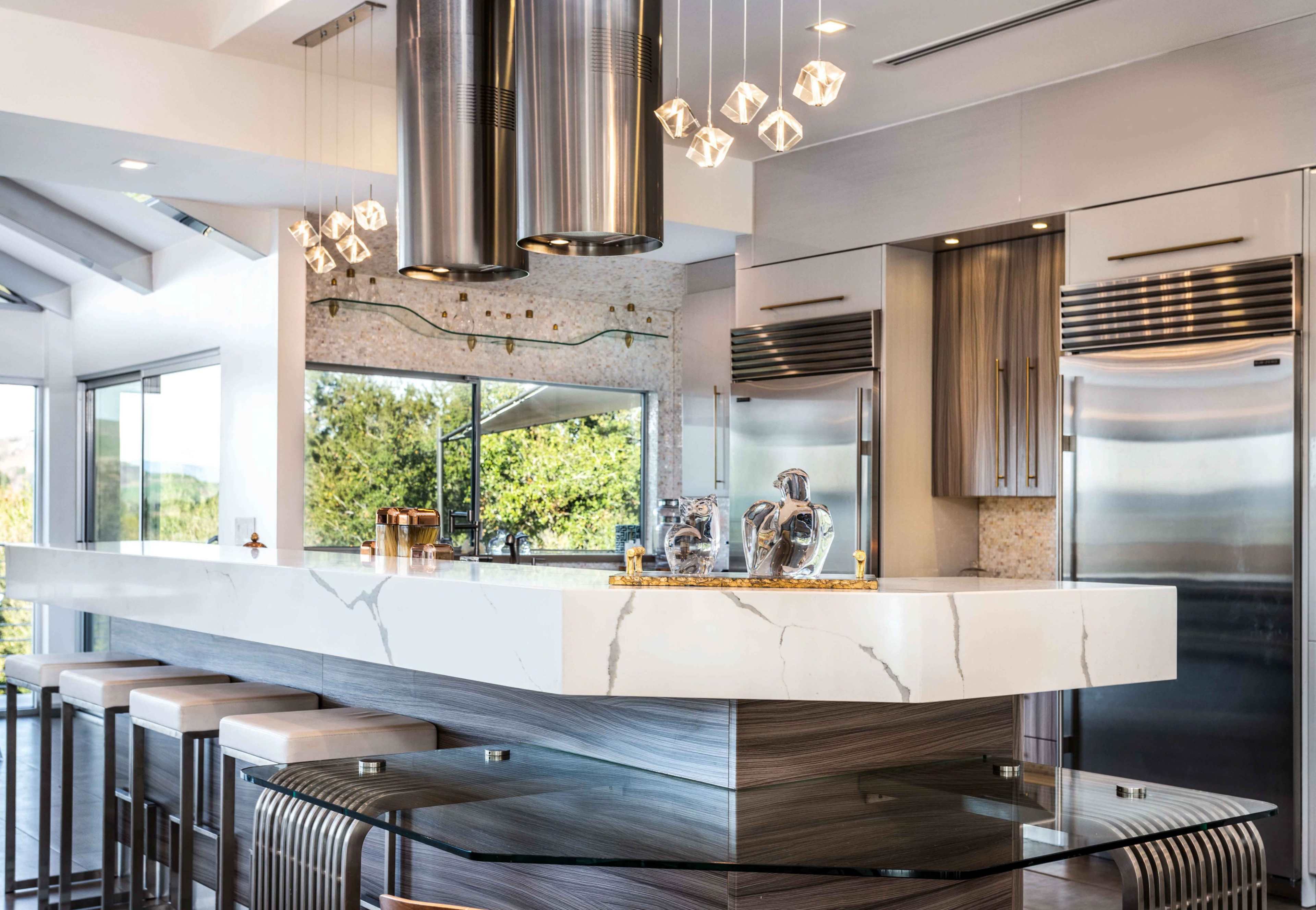A modern kitchen features a large white marble countertop with metallic bar stools and stainless steel appliances, surrounded by ample natural light from large windows.