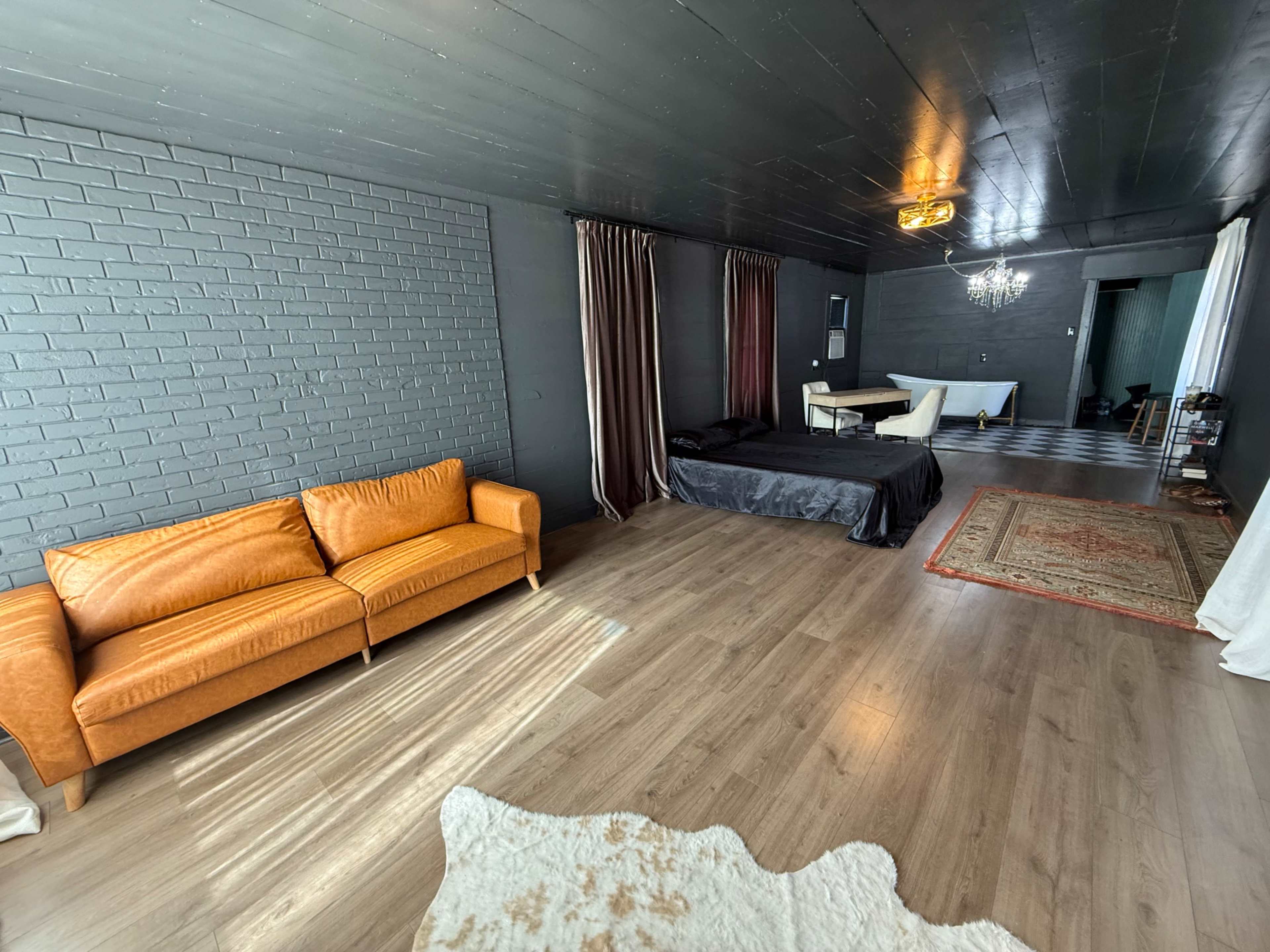 The room features dark walls, a wooden floor, and a mix of furniture including an orange sofa, a black bedspread, and a patterned rug.