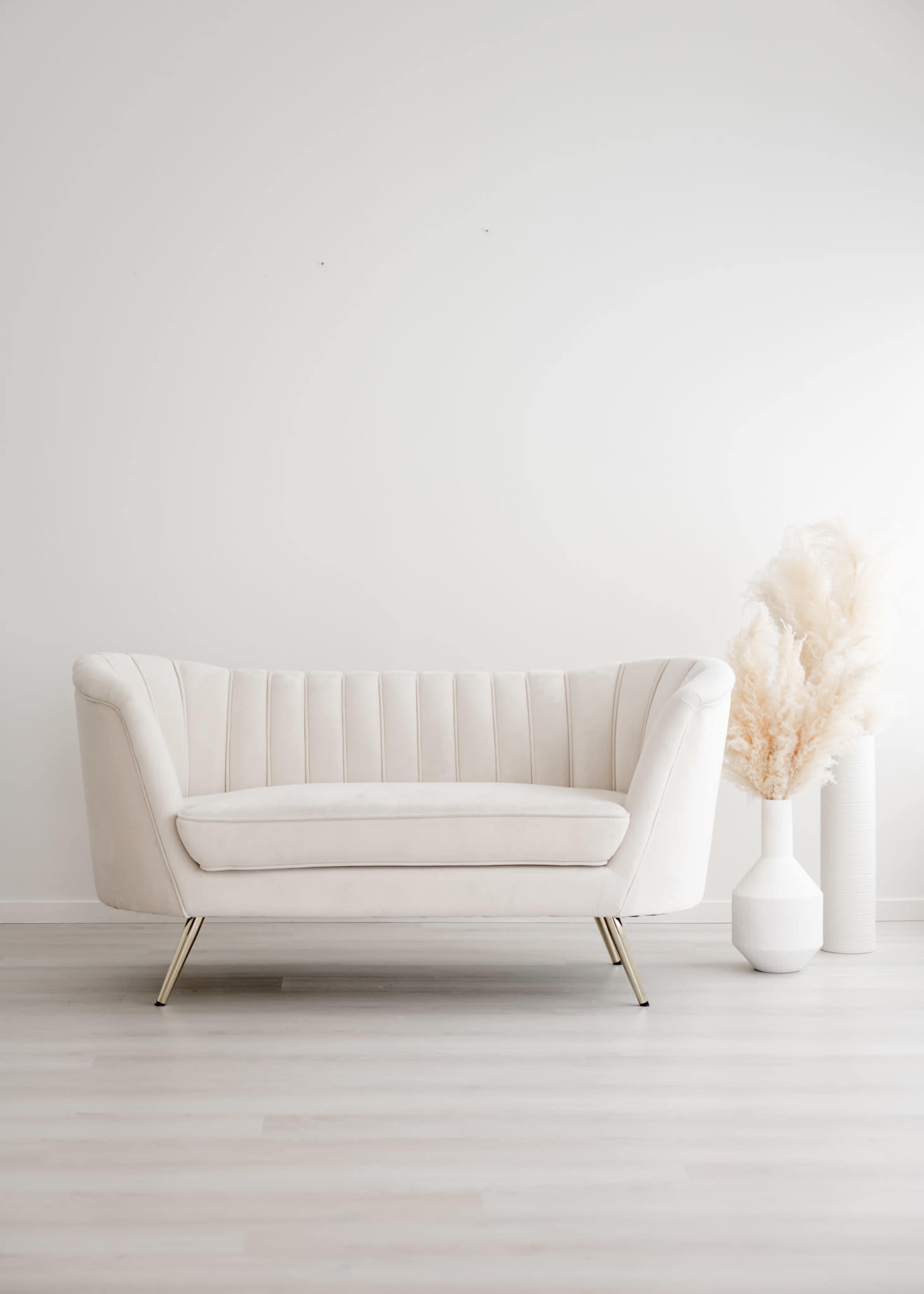 A white, upholstered sofa with gold legs is positioned next to two decorative vases, one white and one filled with dried pampas grass.