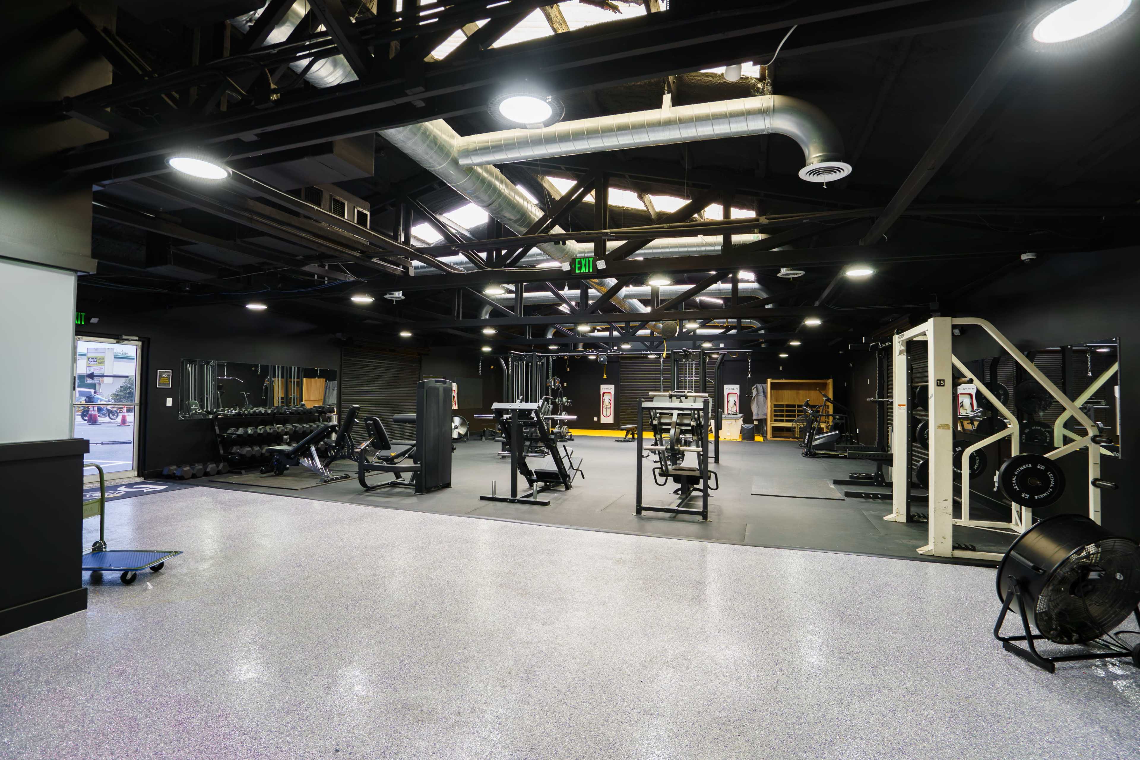 The image shows a spacious gym featuring various workout equipment, including weightlifting machines and free weights, under a modern industrial-style ceiling.