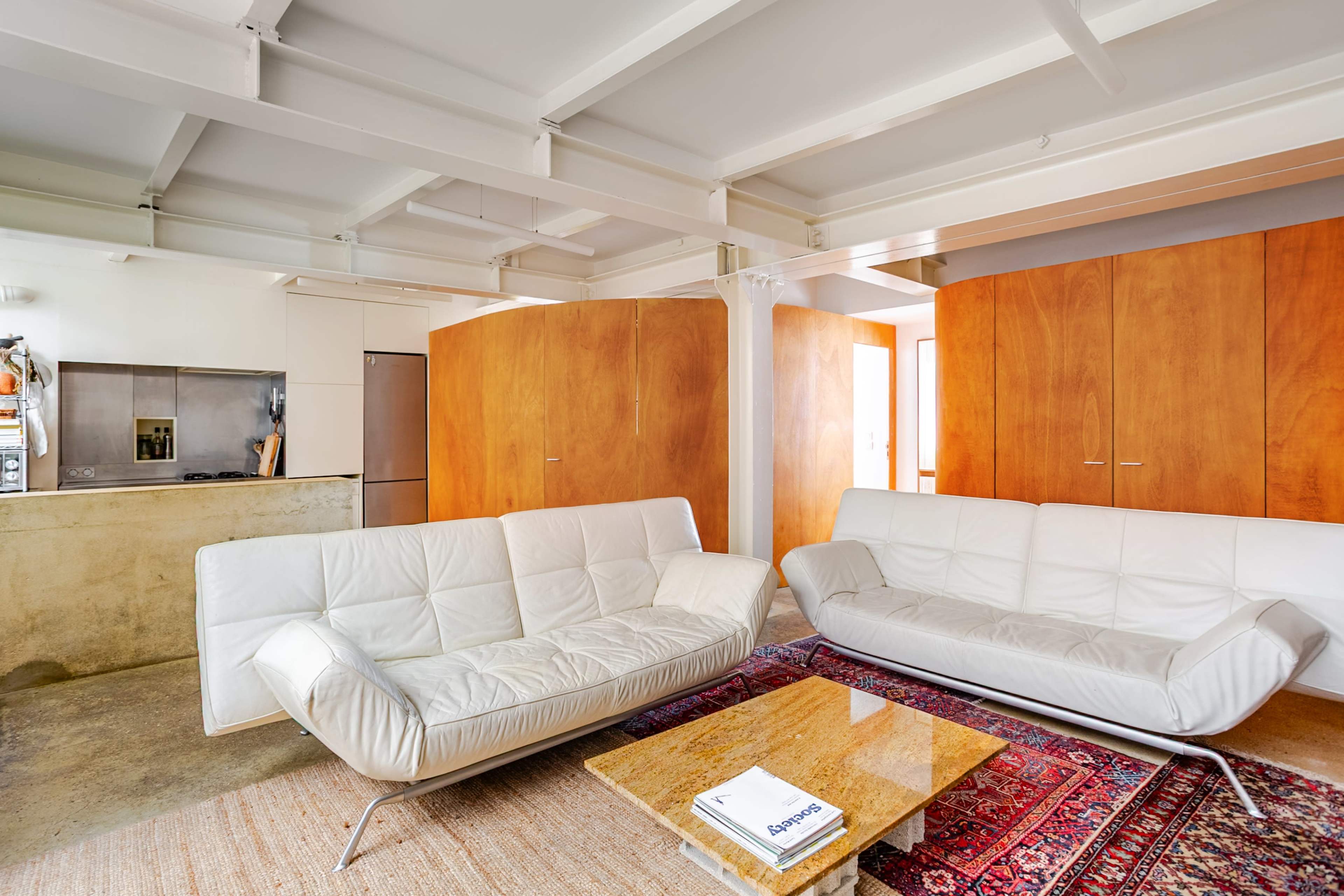 The image depicts a modern living room with white leather sofas, a wooden coffee table, and a minimalist kitchen area visible in the background.