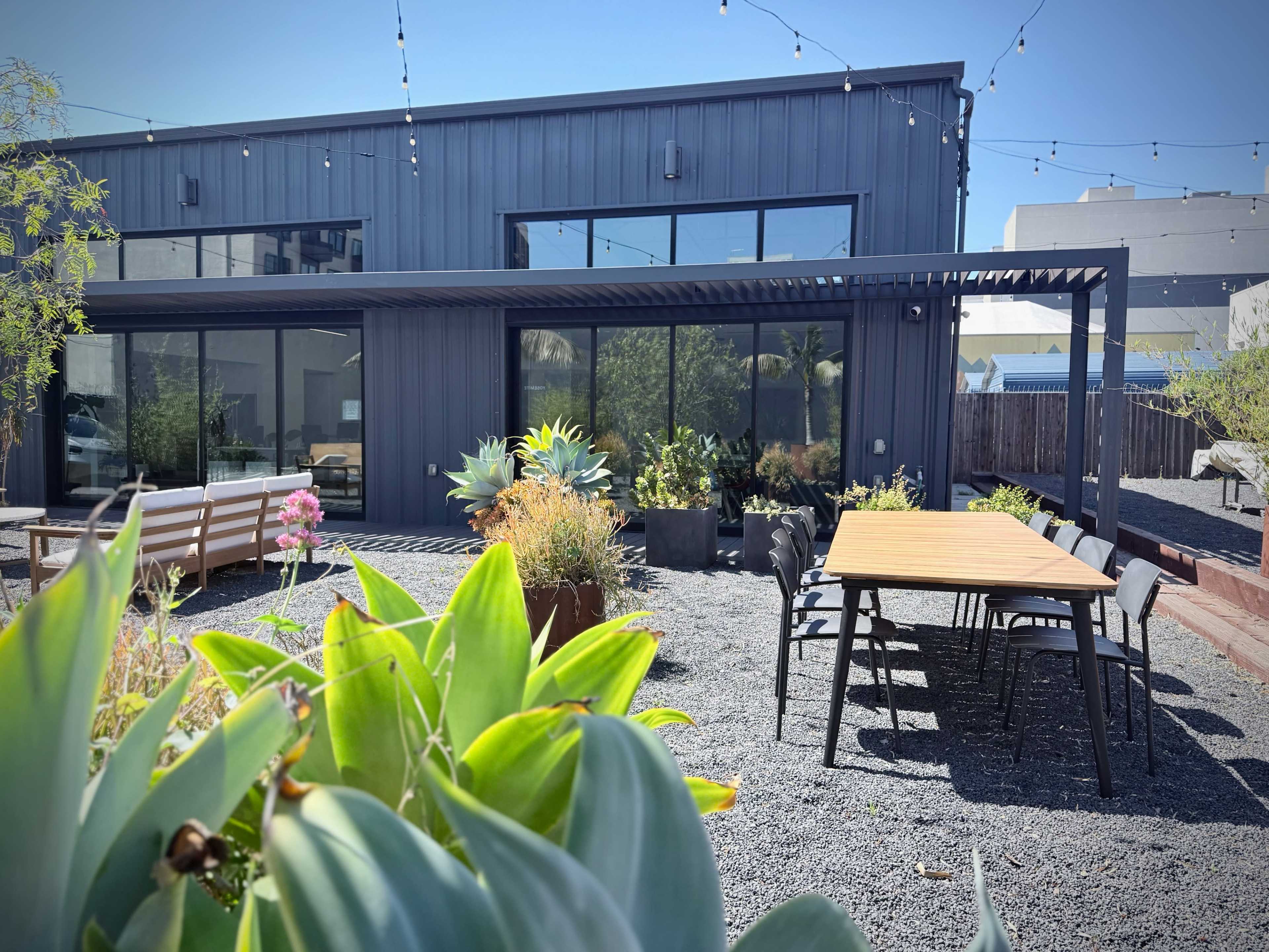 The image shows a modern building with a covered patio area and a landscaped garden featuring succulents and outdoor seating.