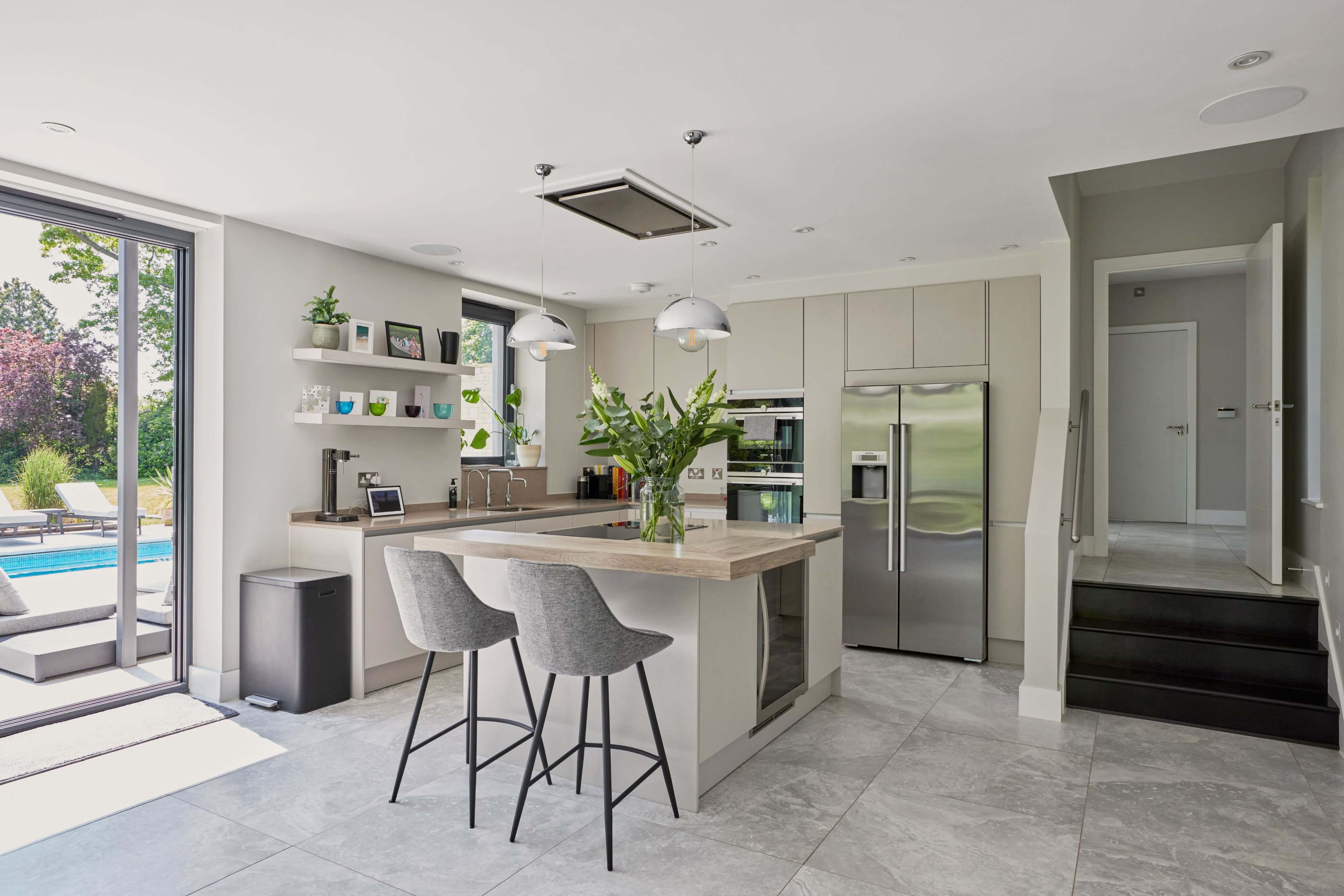 The image shows a modern kitchen with a light-colored island, two bar stools, stainless steel appliances, and a door leading to an outdoor area with a pool.
