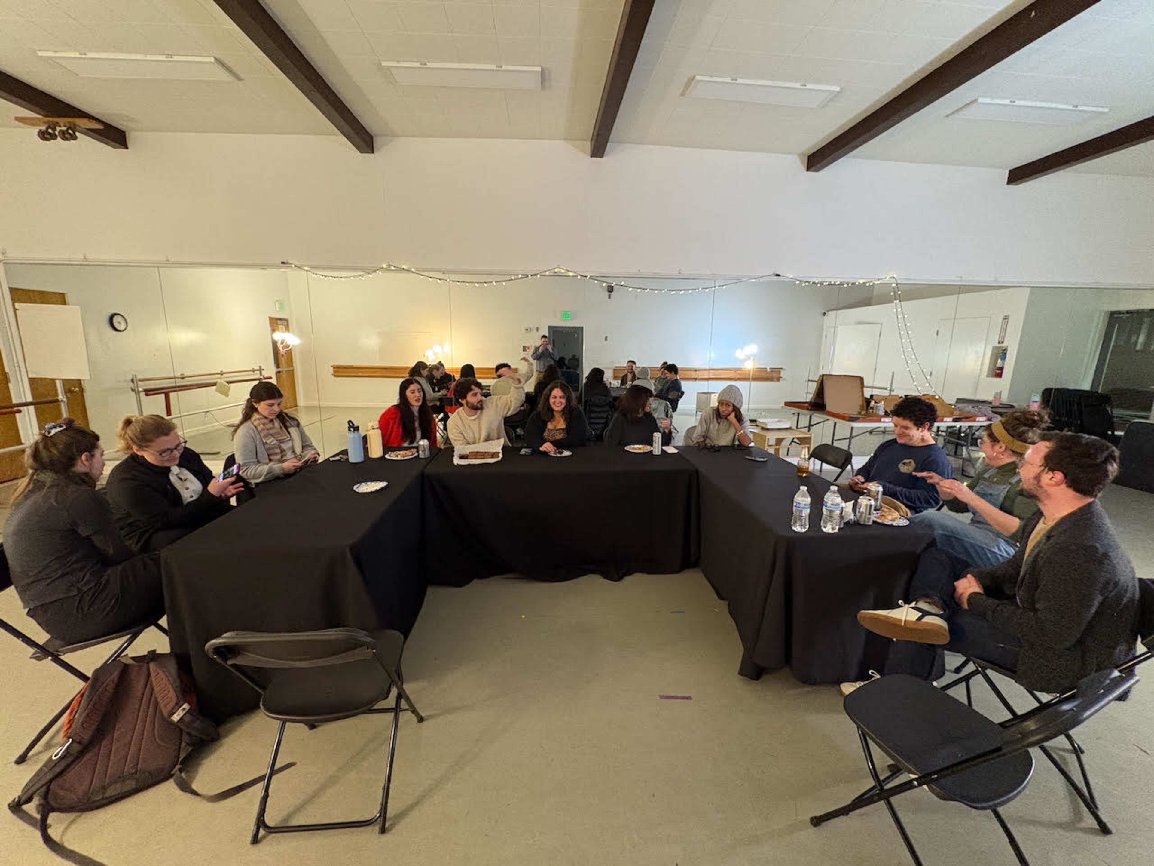 A group of people sits around a large table in a well-lit room, engaged in conversation and eating snacks.