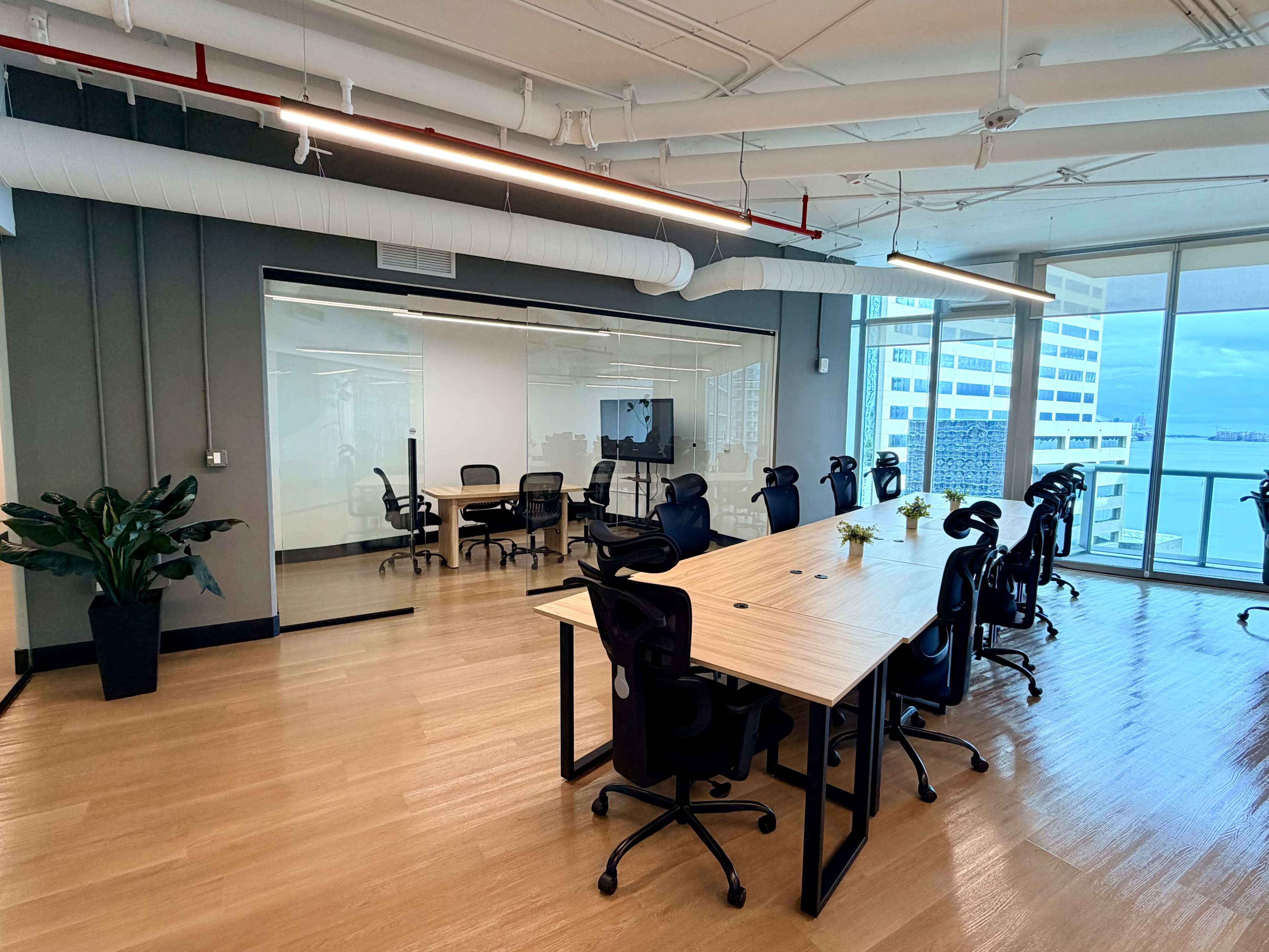 The image shows a modern office space with a large wooden table at the center, surrounded by ergonomic chairs, glass-walled conference rooms, and large windows overlooking a cityscape.