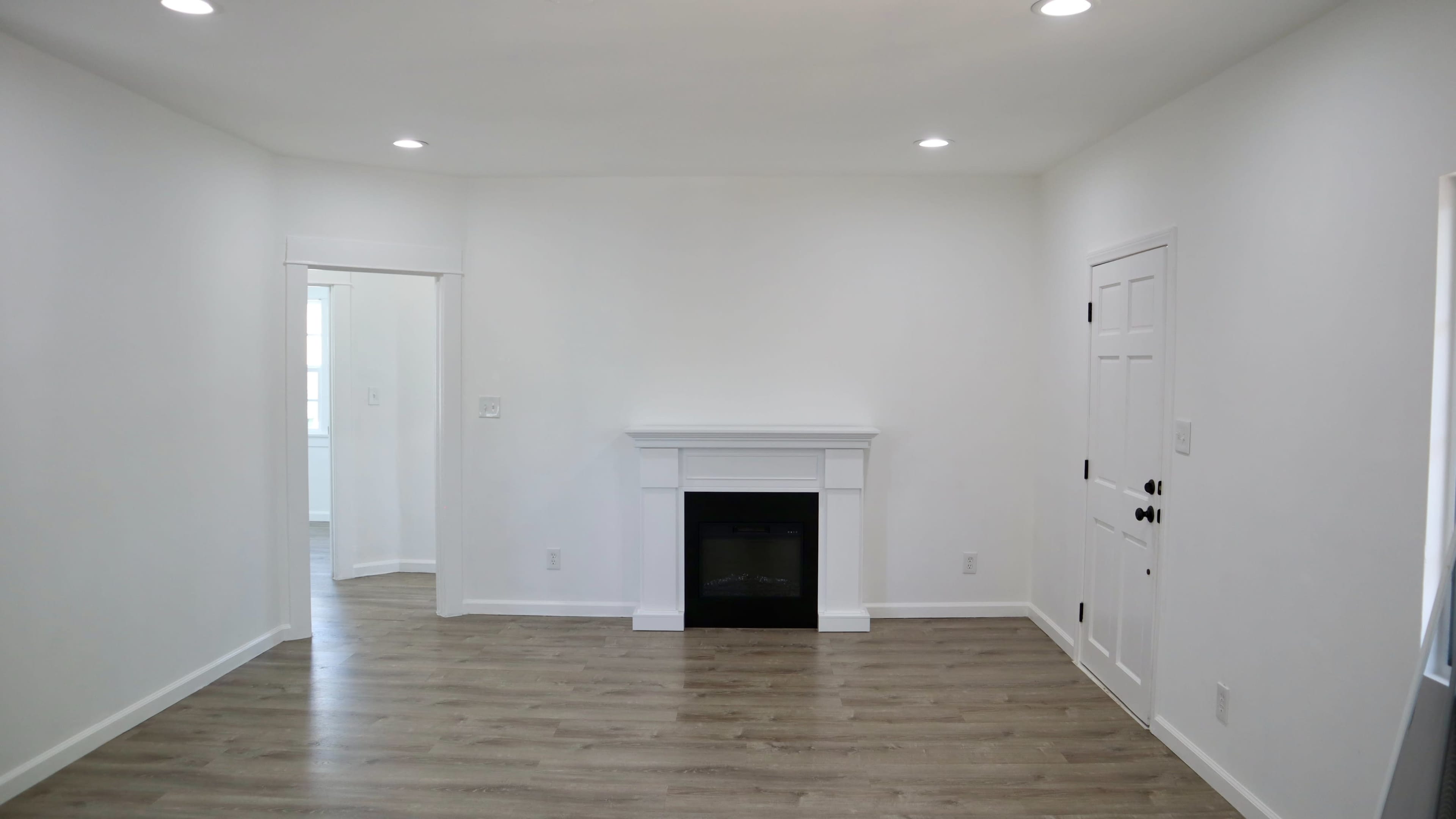 A well-lit room with a white fireplace, light wood flooring, and an open doorway leading to another space.