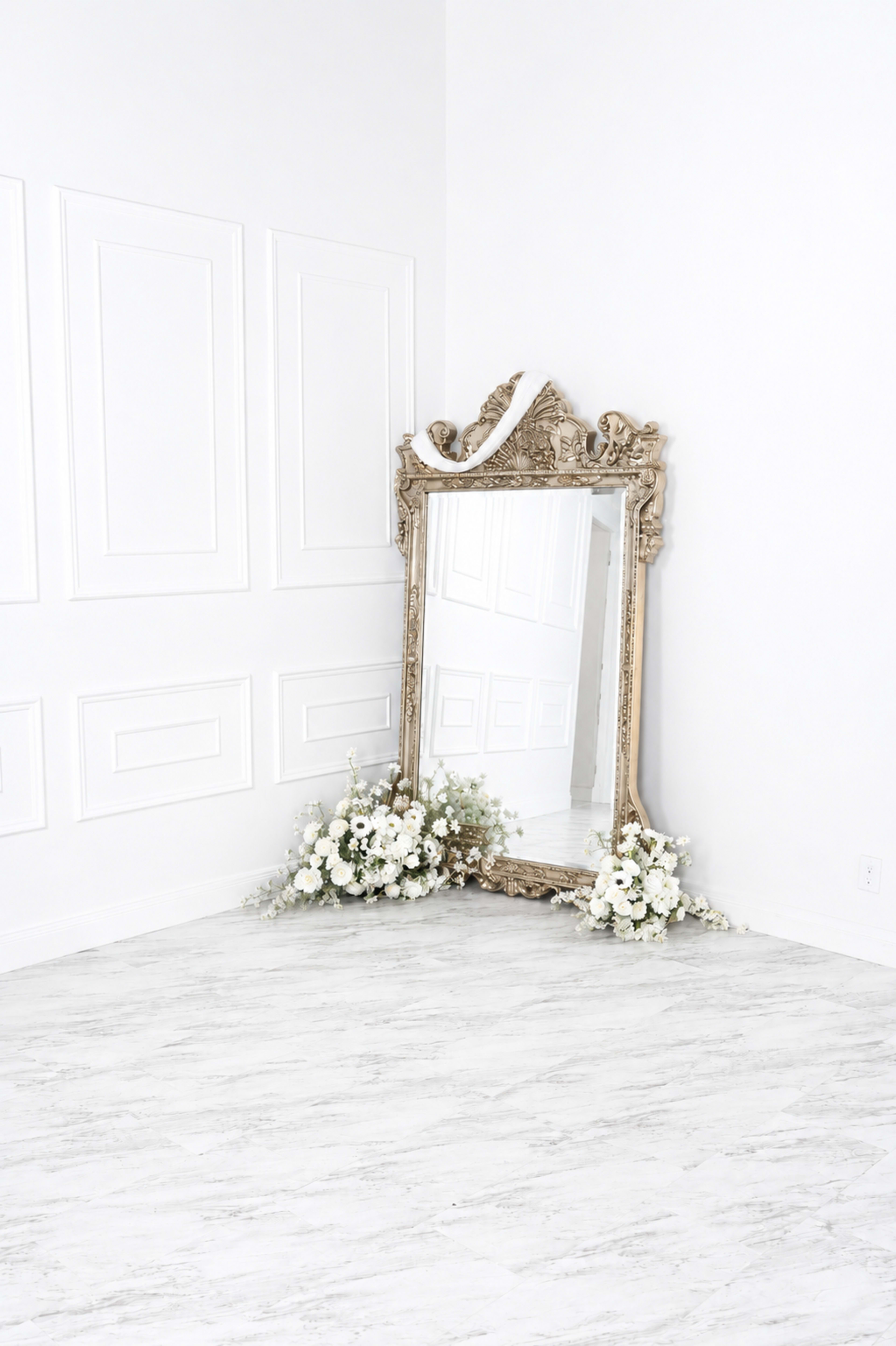 A large, ornate mirror is positioned in a corner of a bright room, surrounded by arrangements of white flowers.