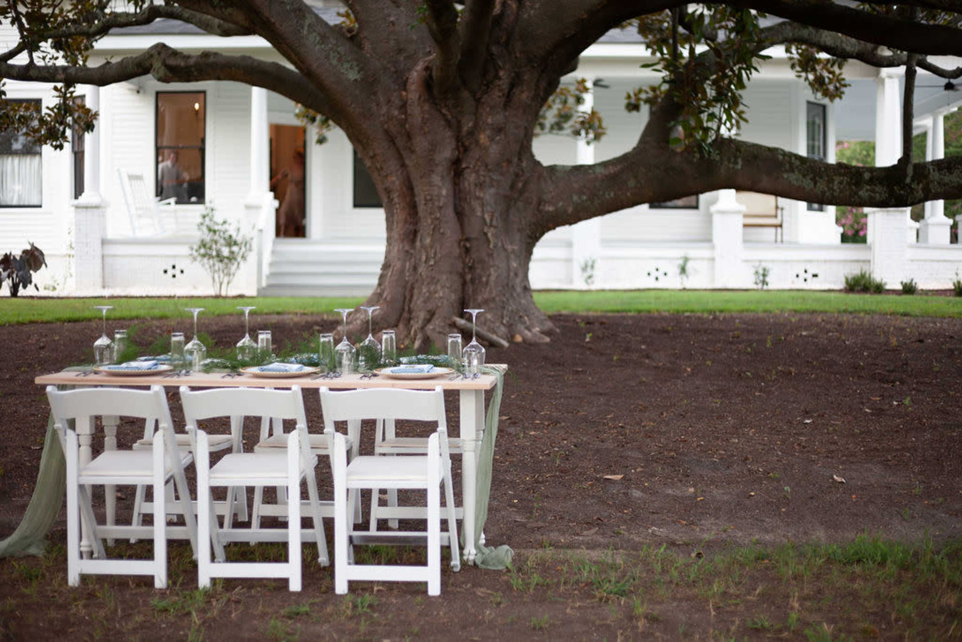 A dining table with white chairs is set under a large tree, near a white house in the background.