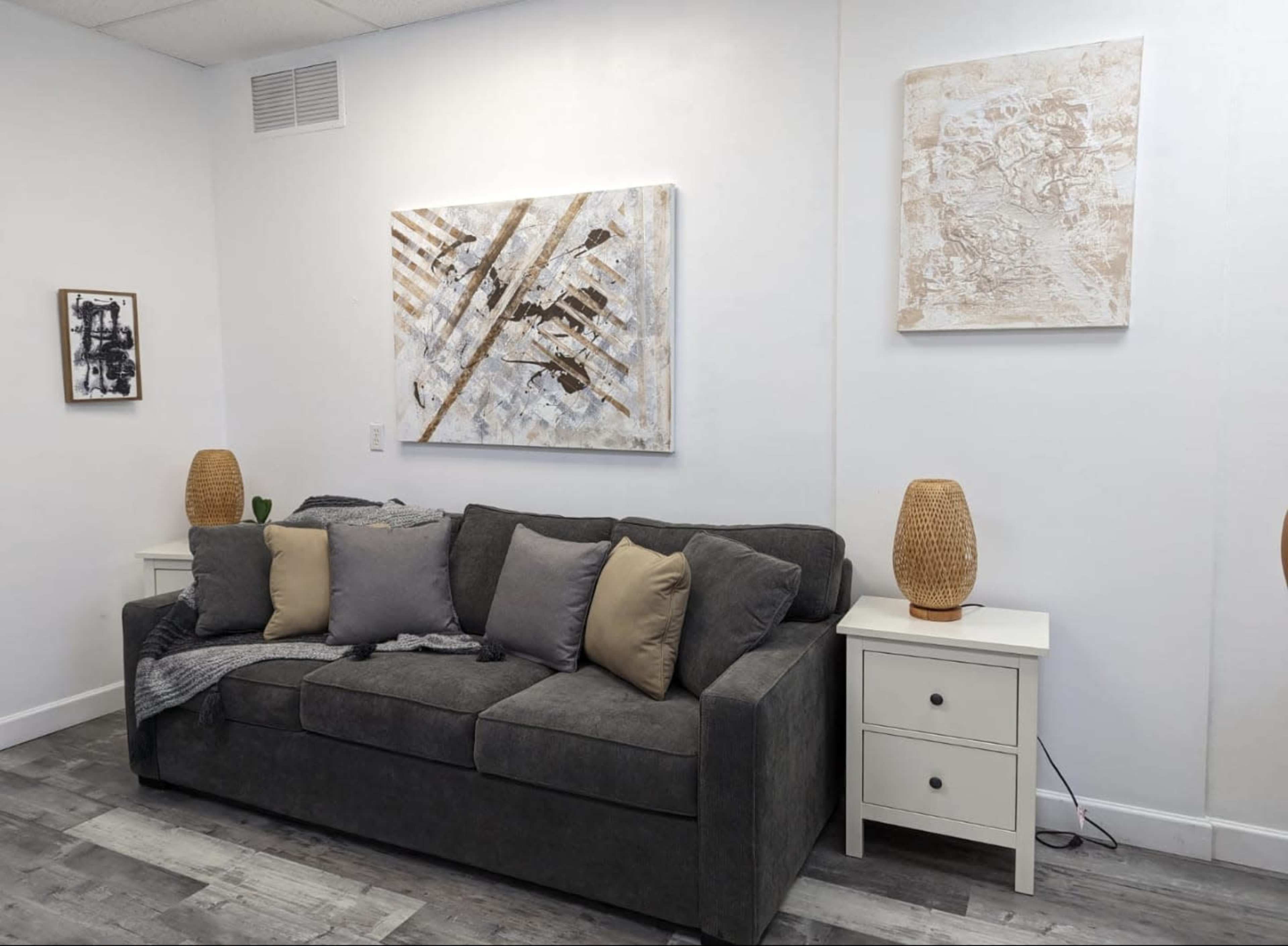 The scene depicts a modern living room with a dark gray sofa adorned with pillows, two light-colored side tables, and abstract artwork on the walls.