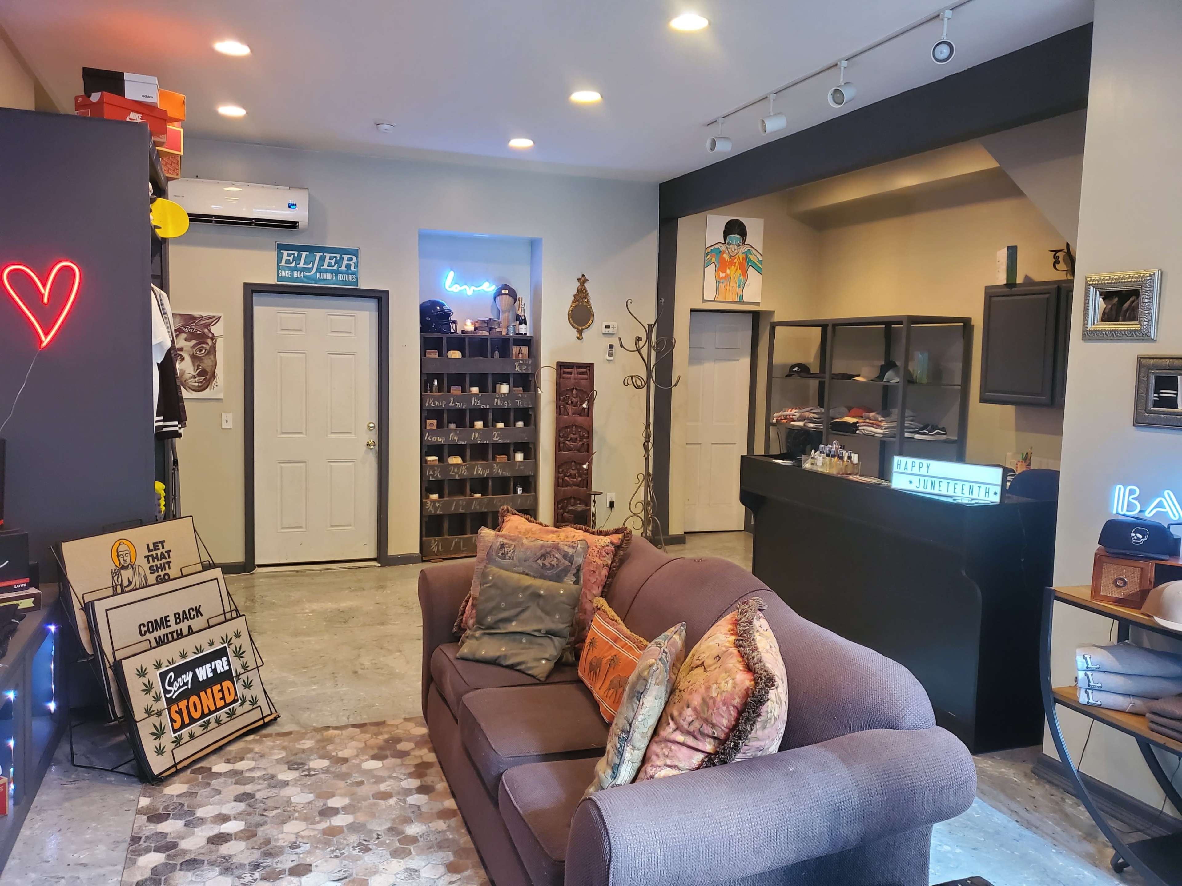 A cozy interior space featuring a sofa with decorative pillows, shelves filled with items, a small service counter, and wall decorations including signs and a neon heart.