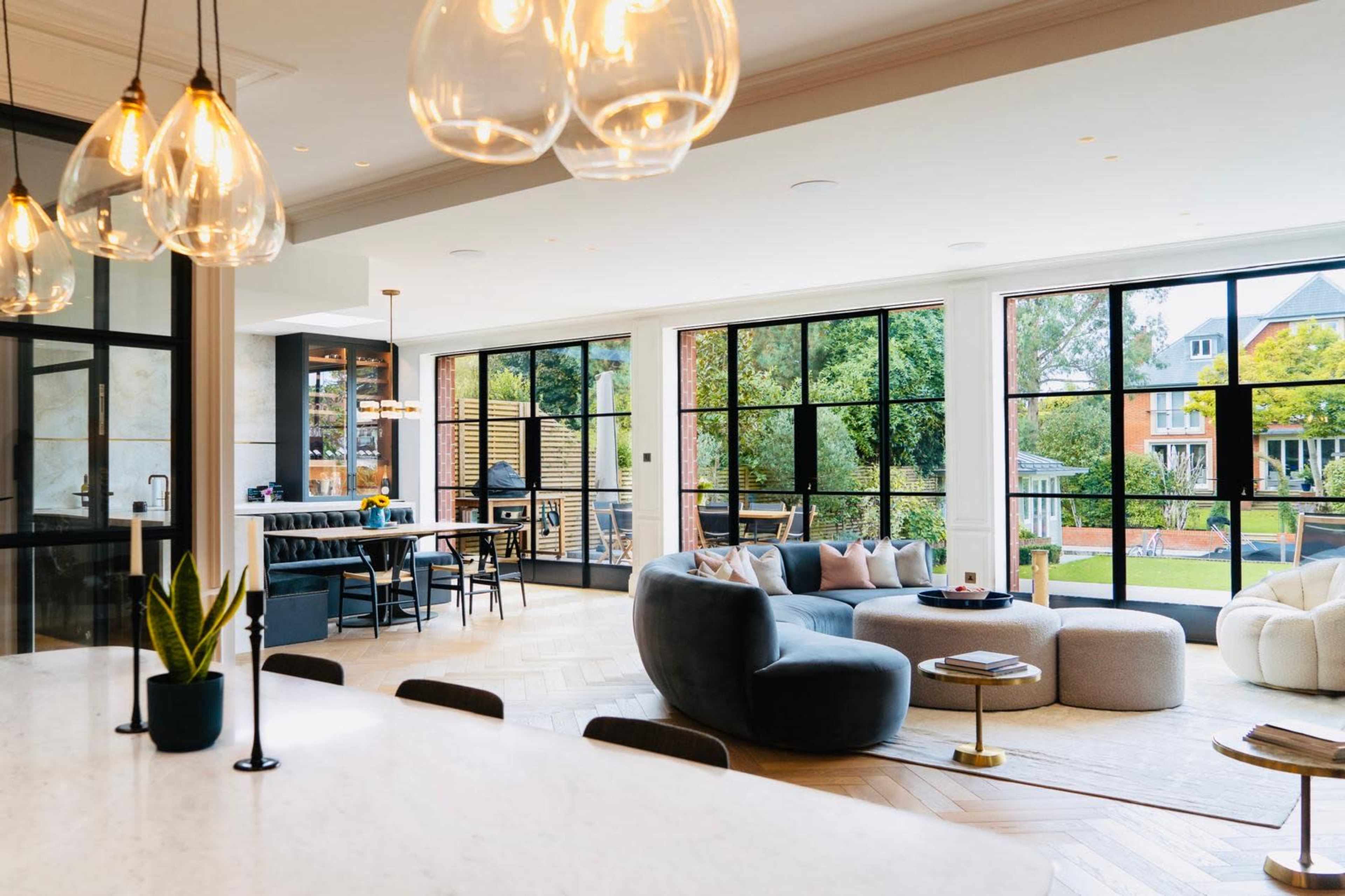 The image depicts a modern open-plan living space featuring a lounge area with a curved sofa, a coffee table, and large windows that overlook a garden, alongside a dining area with a table and chairs.