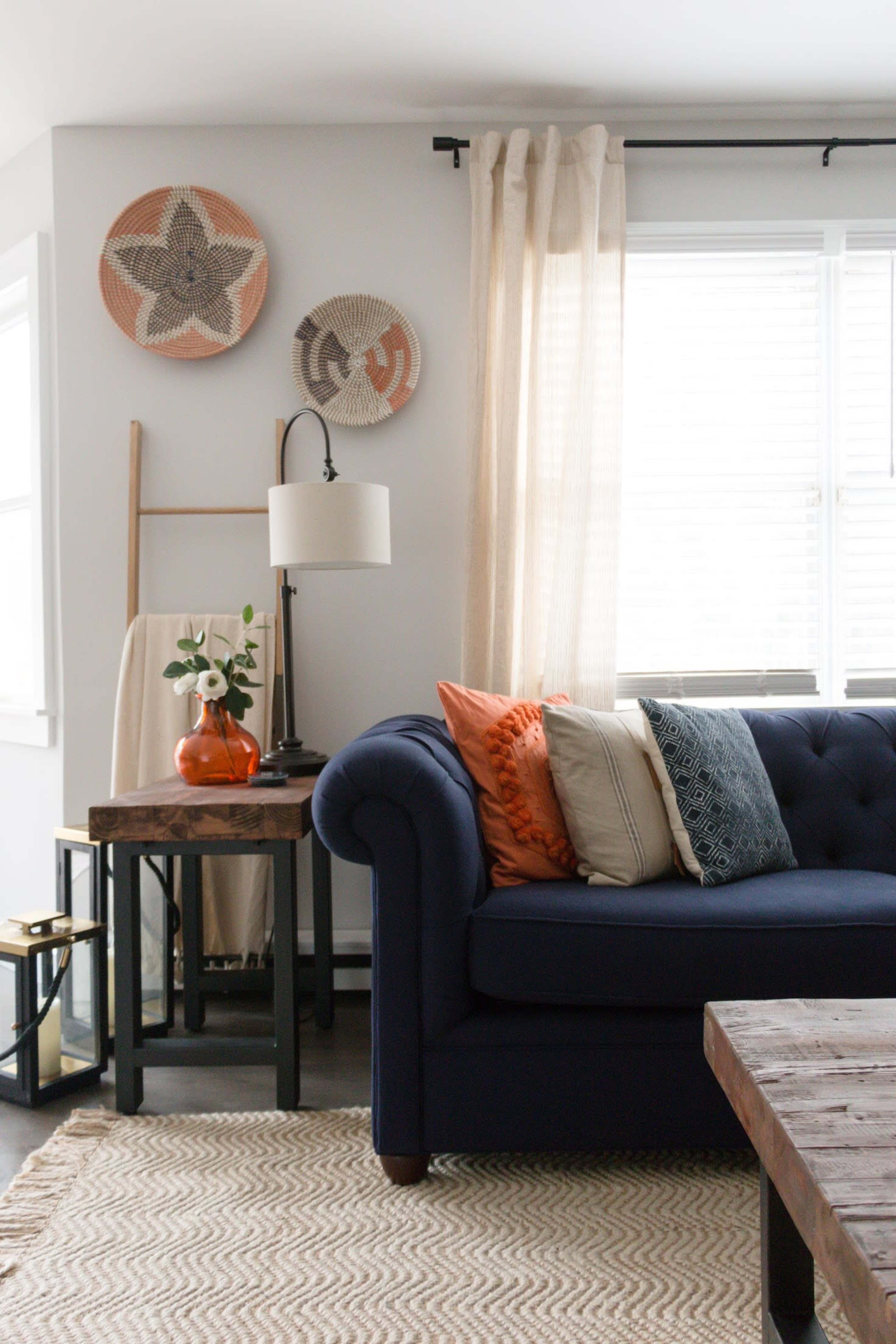 A navy blue sofa with decorative pillows sits beside a wooden table and a lamp, with woven wall art and a sheer curtain in the background.