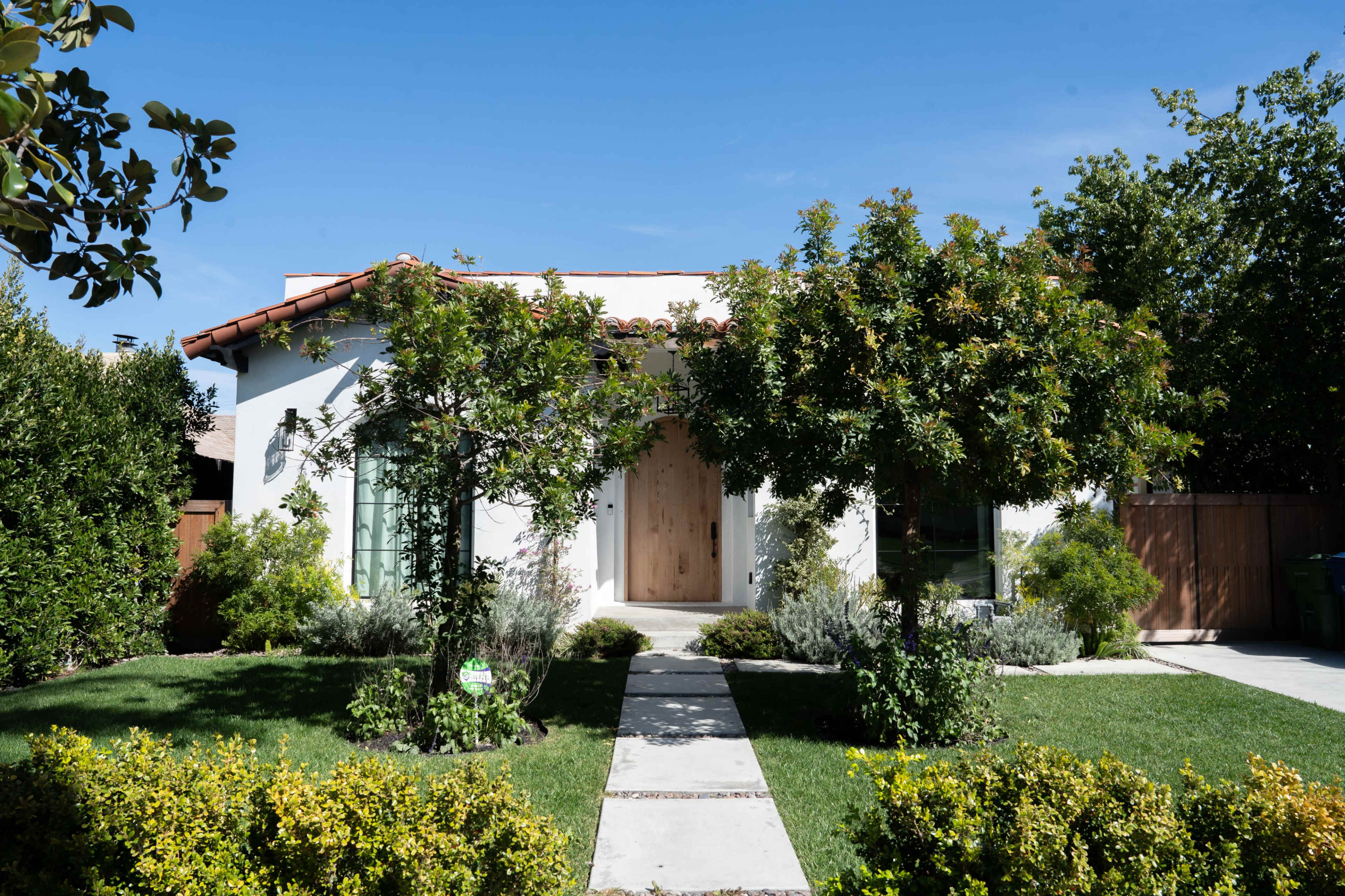 A white house with a wooden door is flanked by trees and surrounded by a well-maintained garden and pathway.