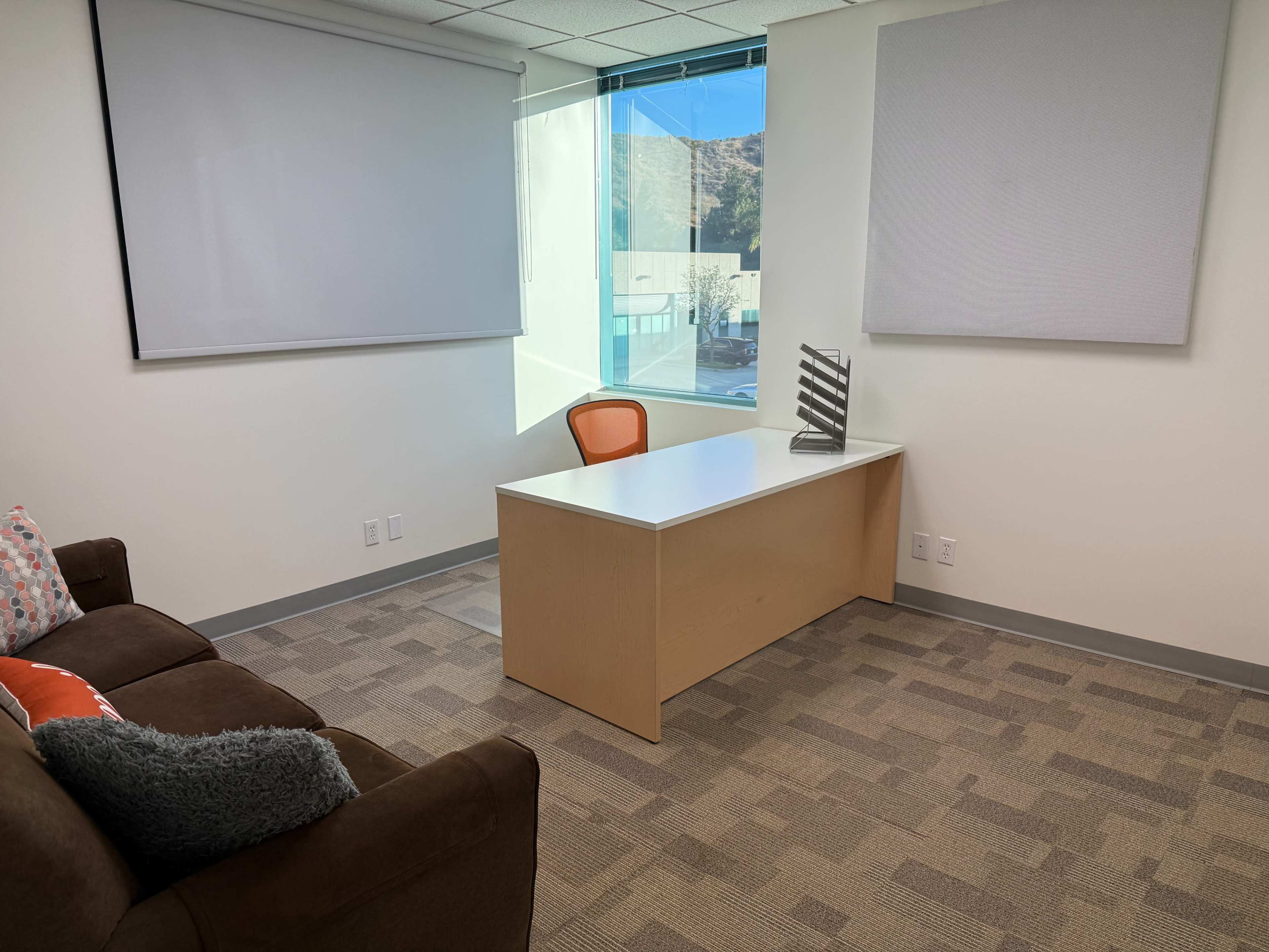 The image shows a modern office space featuring a desk, an orange chair, a brown sofa, and a large window with natural light.