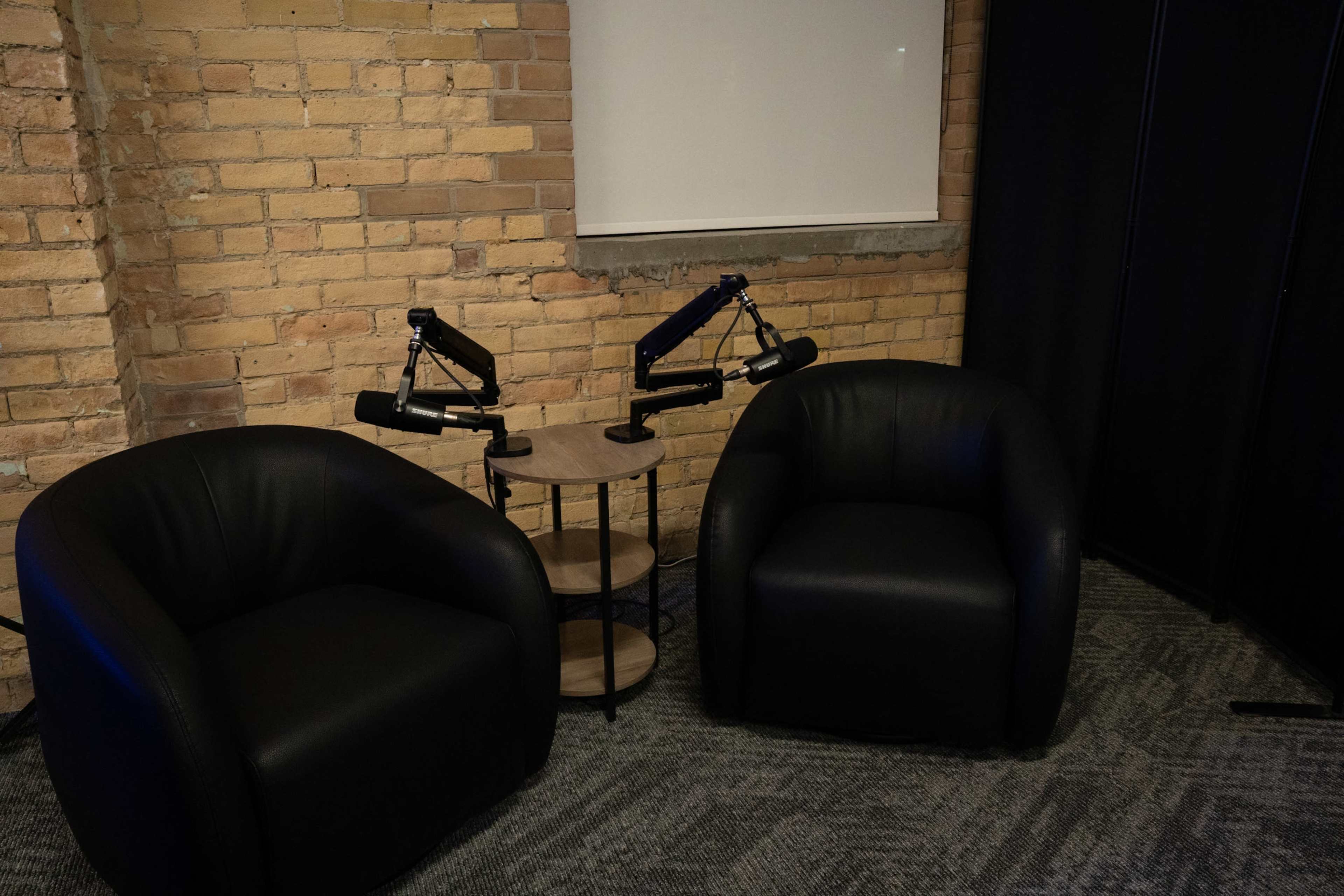 The image shows two black upholstered chairs placed opposite each other beside a small circular table with two mounted microphones in a room with exposed brick walls.