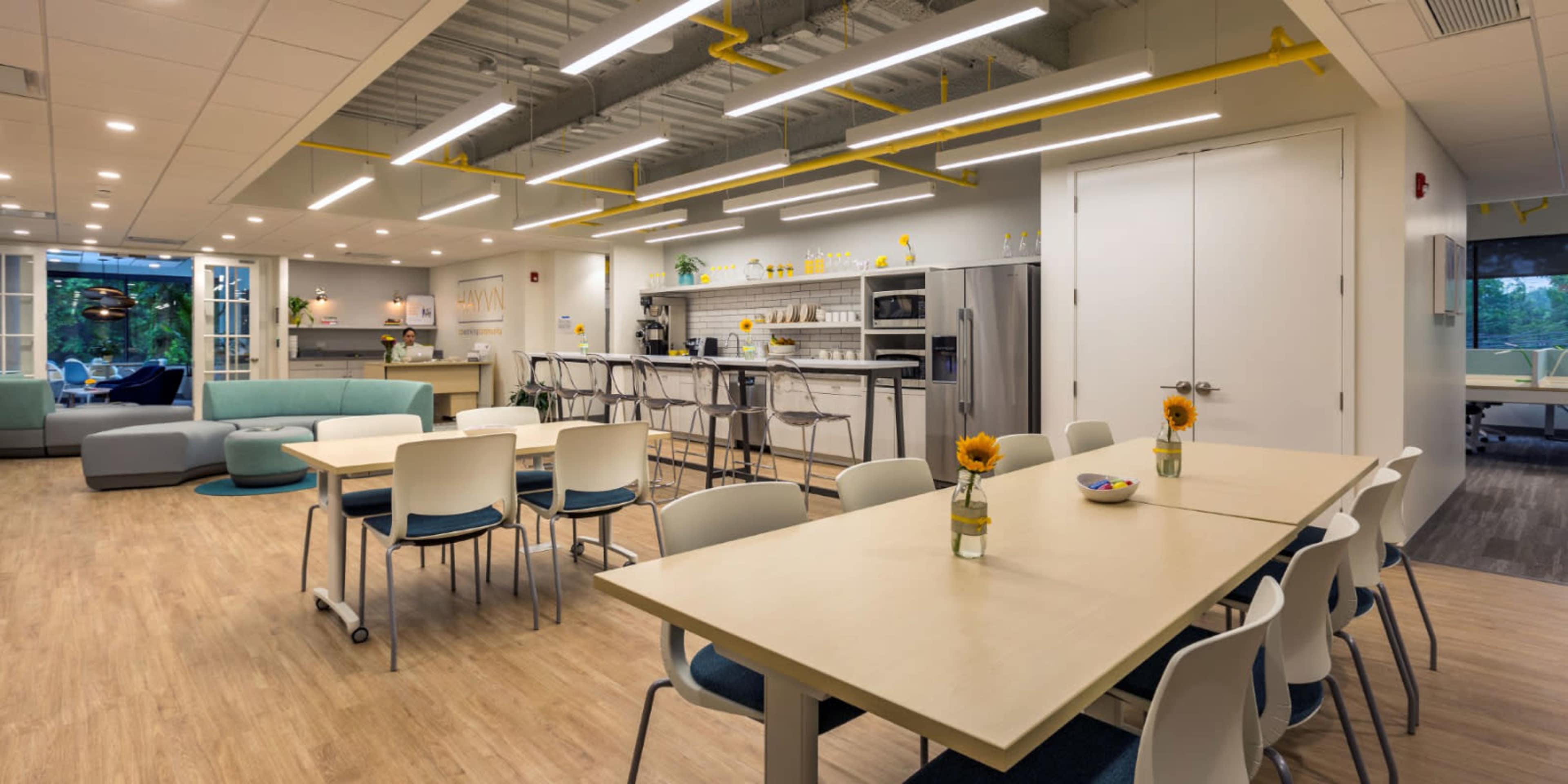 A spacious, modern break room features light wood flooring, multiple tables, a kitchenette with stainless steel appliances, and decor including sunflowers.