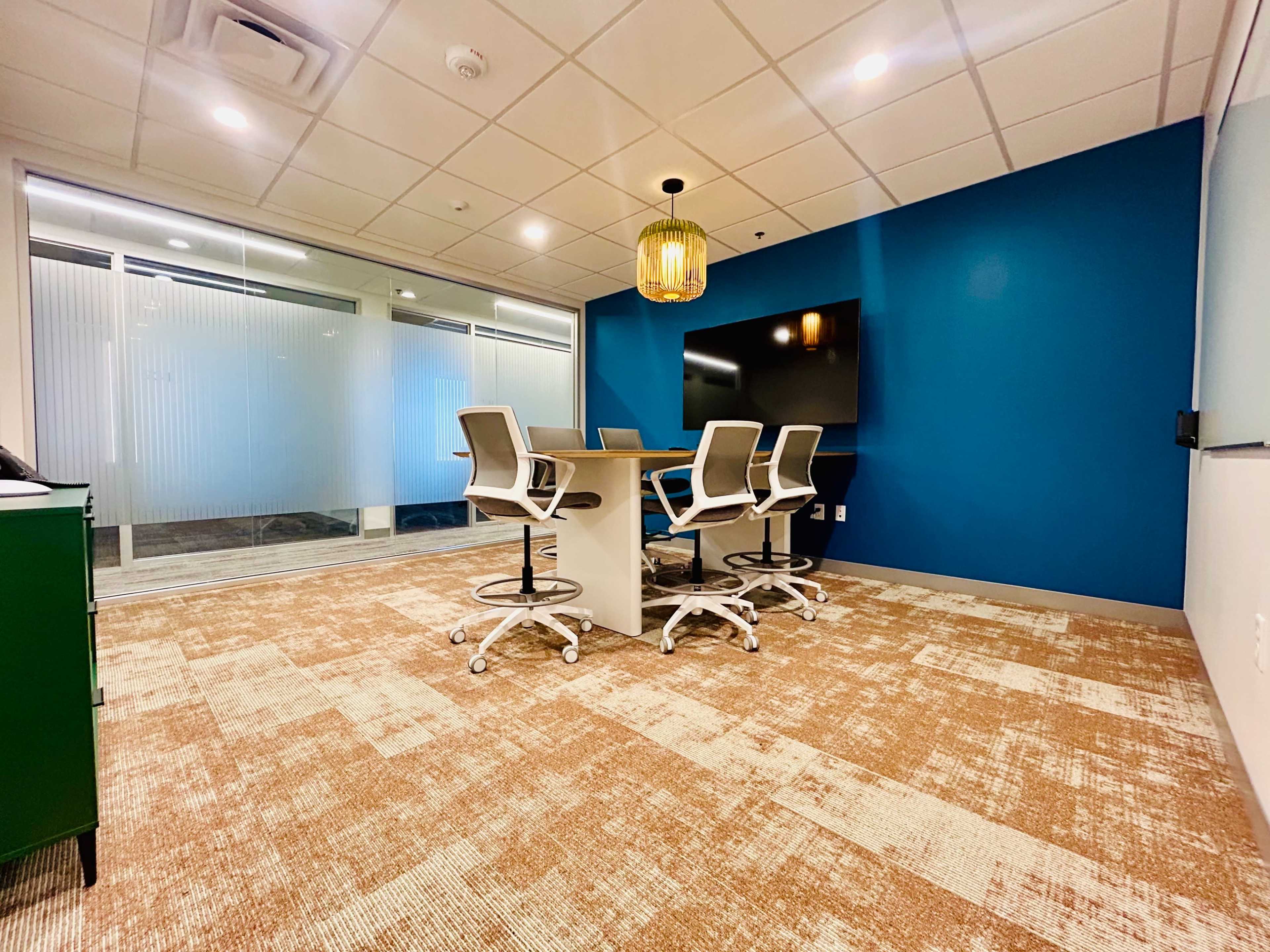 The image shows a modern conference room with a circular table, four ergonomic chairs, and a large screen mounted on a blue accent wall.