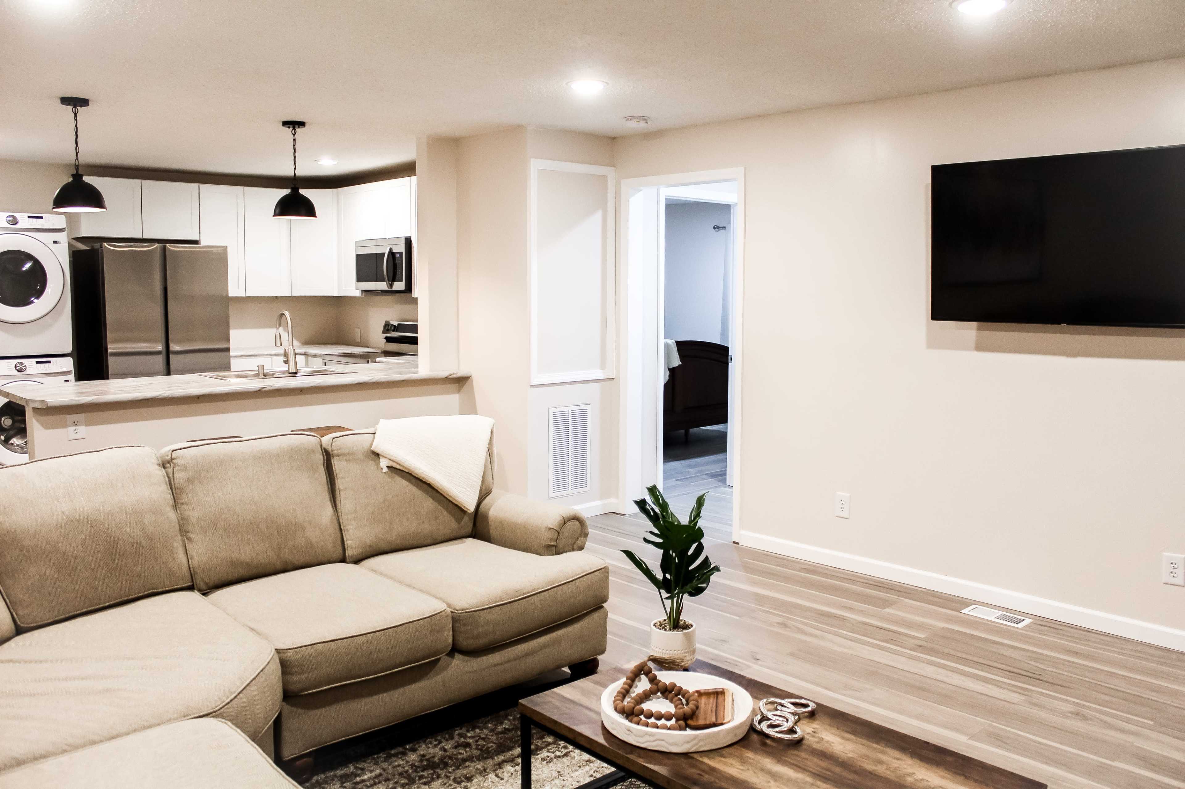 The image shows a modern living room with a beige sectional sofa, a wooden coffee table, a wall-mounted TV, and an open kitchen area with white cabinets in the background.