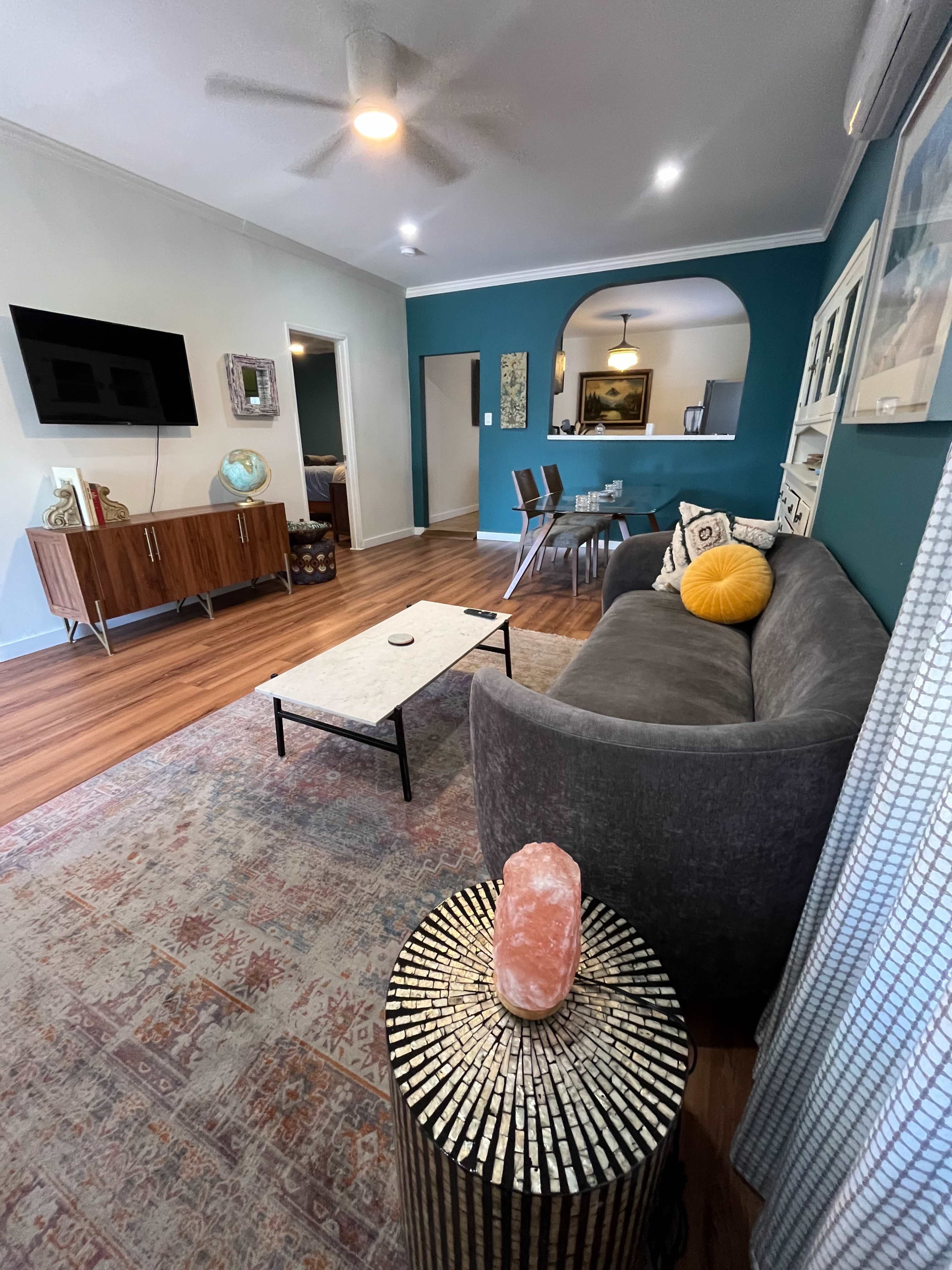 The image shows a living room with a gray sofa, a coffee table, a TV mounted on the wall, and a dining area in the background decorated with a large artwork.