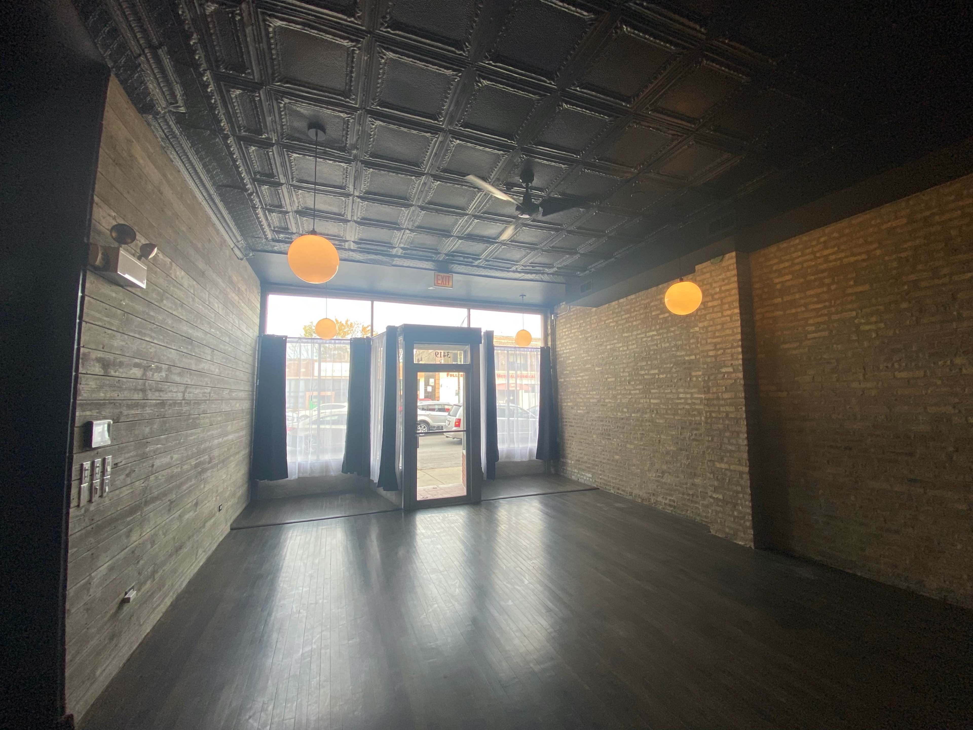 An empty room with wooden walls, large windows at the far end, and two doors leading outside, illuminated by pendant lights.