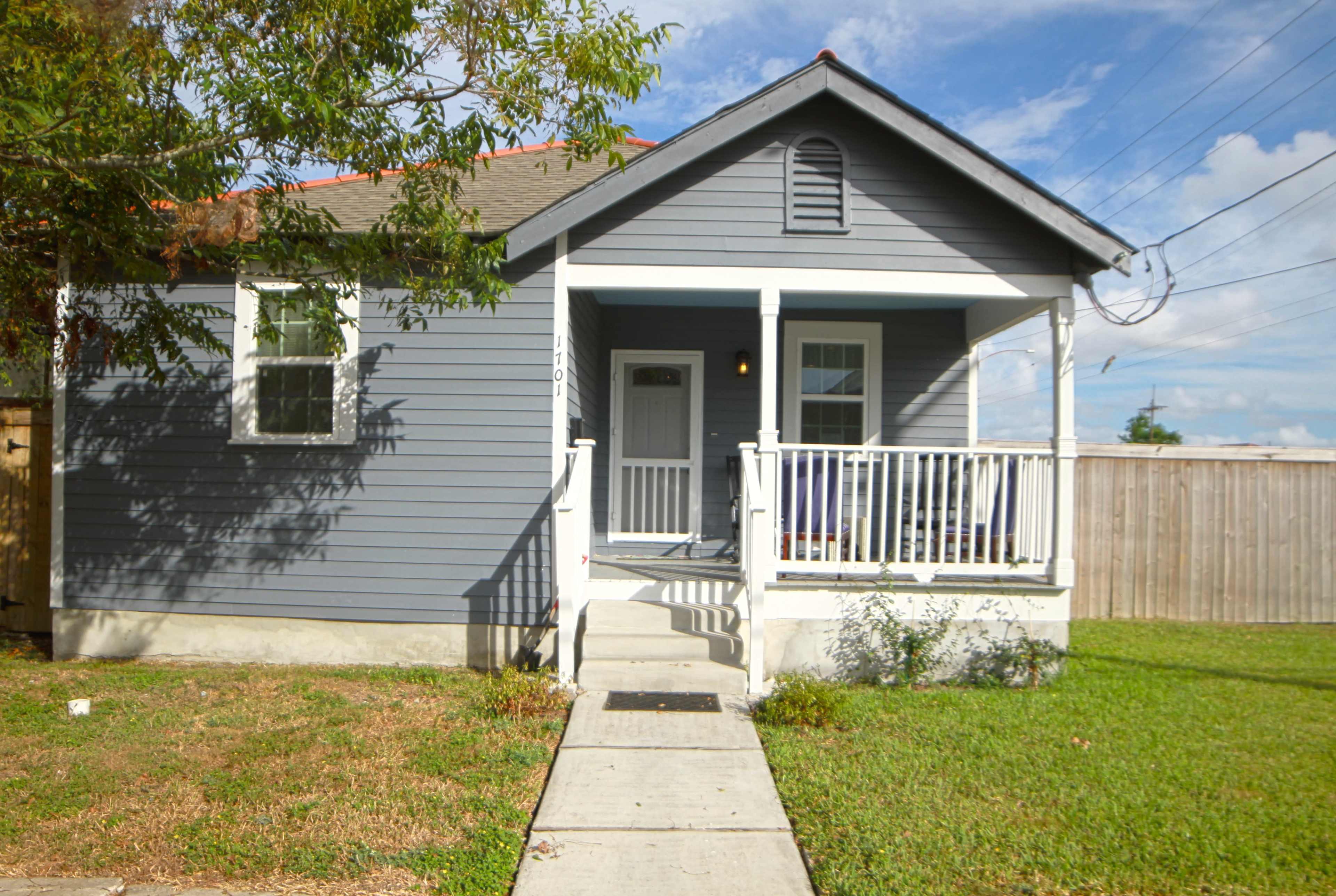 A gray house with a front porch and white railings is situated on a grassy lawn, with a concrete walkway leading to the entrance.