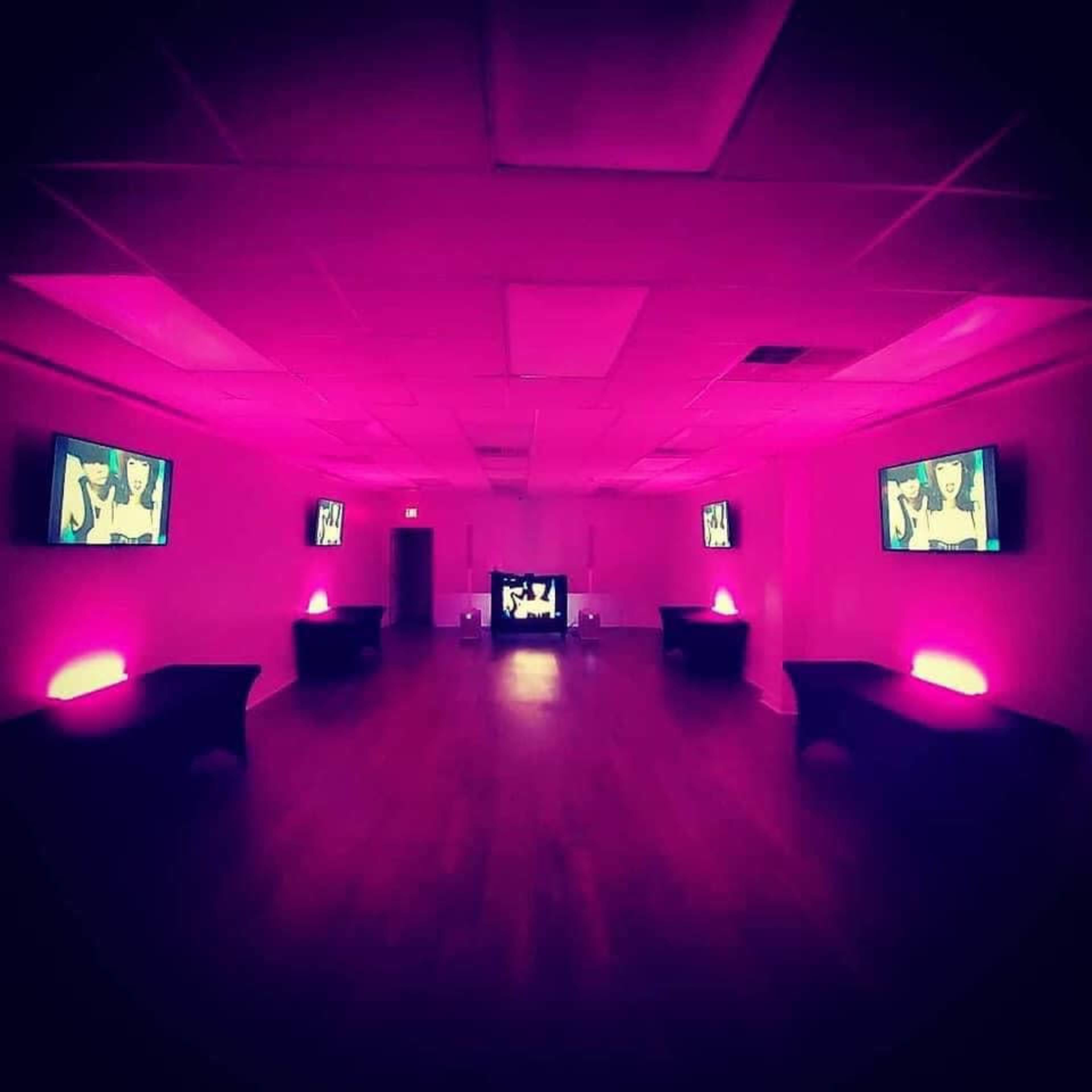 A room illuminated in pink light, featuring multiple screens displaying images on the walls and a central television.
