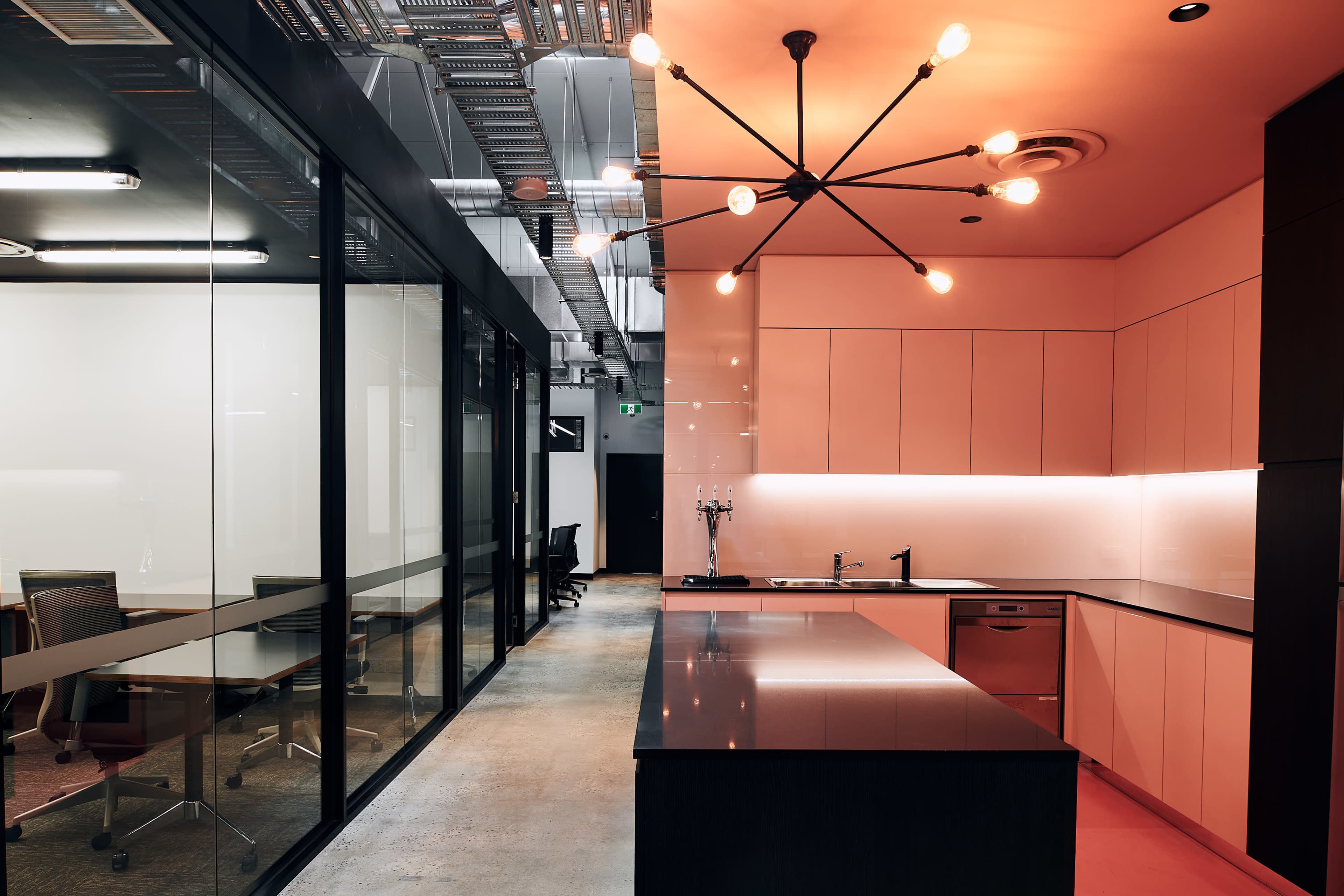 The image shows a modern office kitchen area featuring a central island, a minimalist design with pink cabinetry, and a lighting fixture with multiple bulbs.