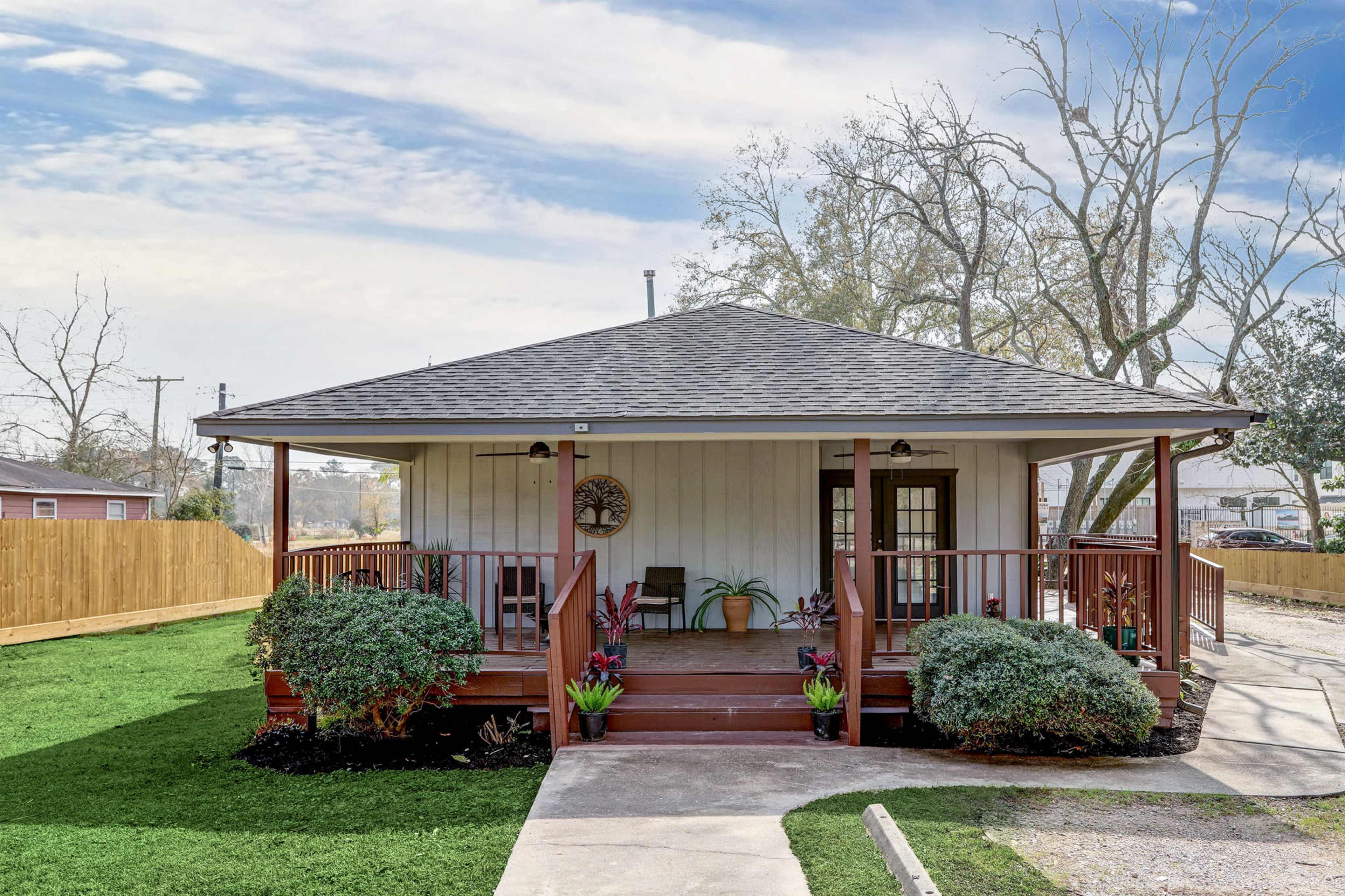 A single-story house with a triangular roof and front porch is surrounded by neatly trimmed grass and small plants.