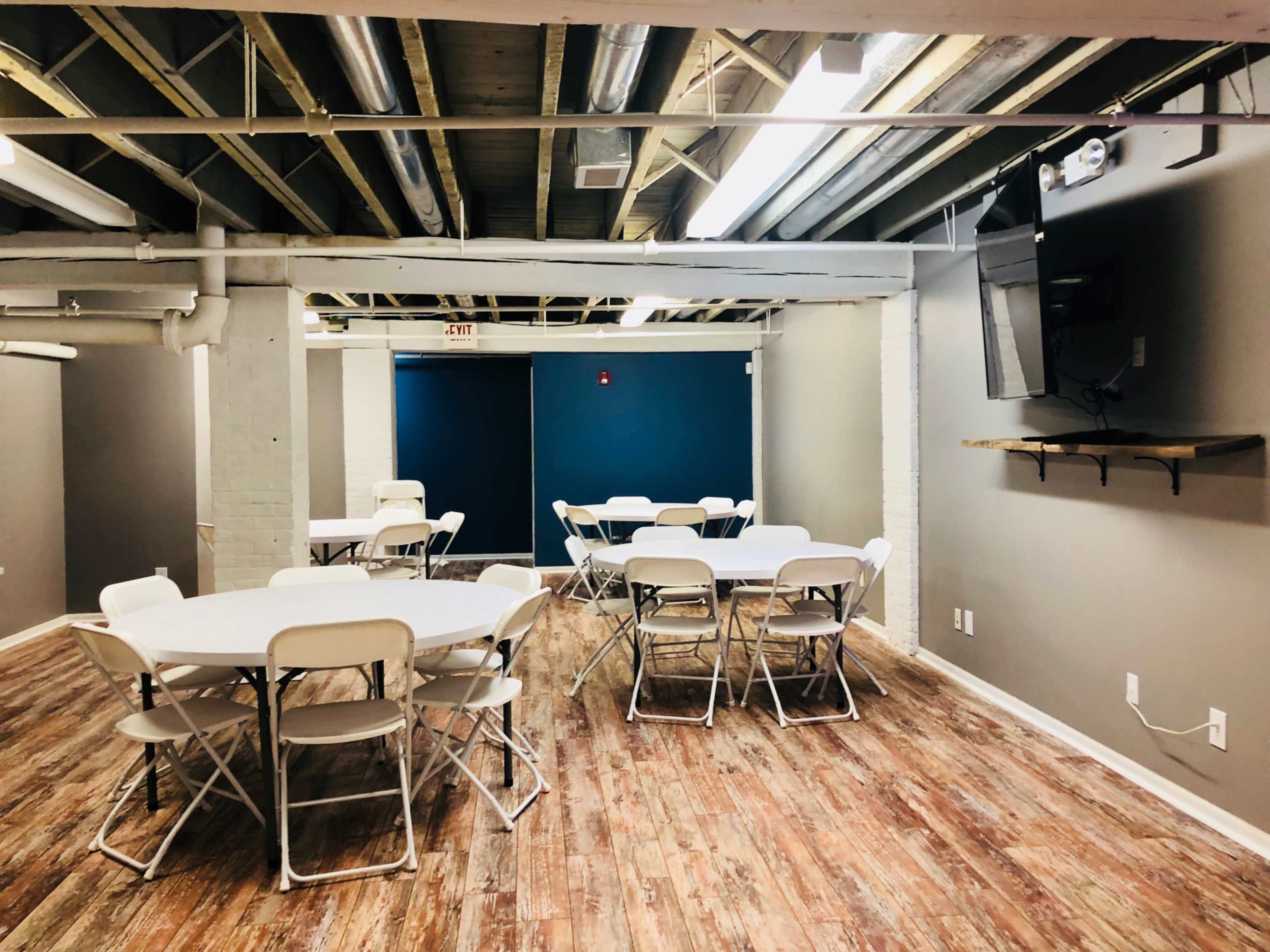 A spacious room with several round tables and chairs, featuring exposed beams and a television mounted on the wall.