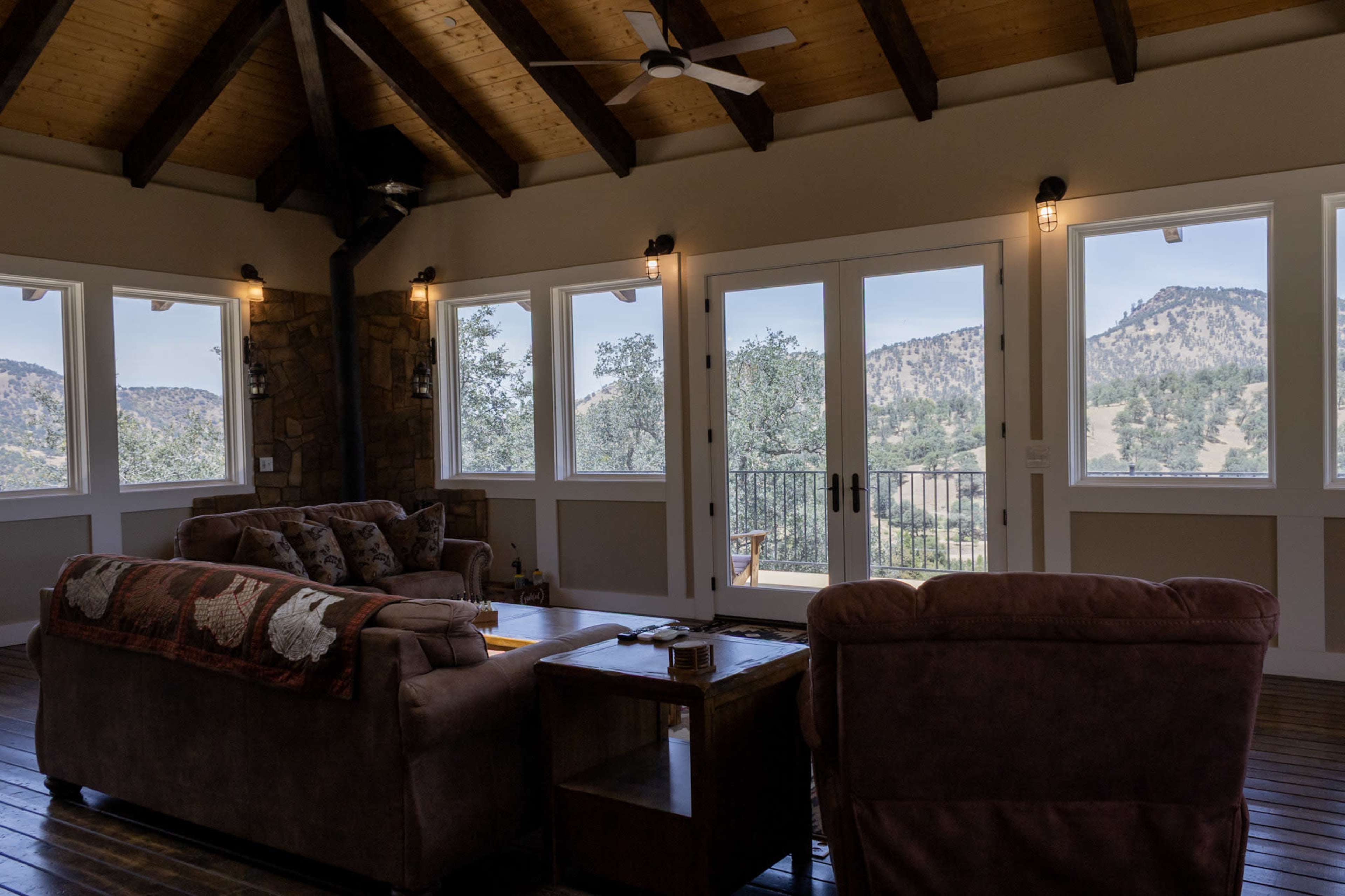 The image shows a living room with a wooden ceiling, two sofas, and large windows overlooking a mountainous landscape.