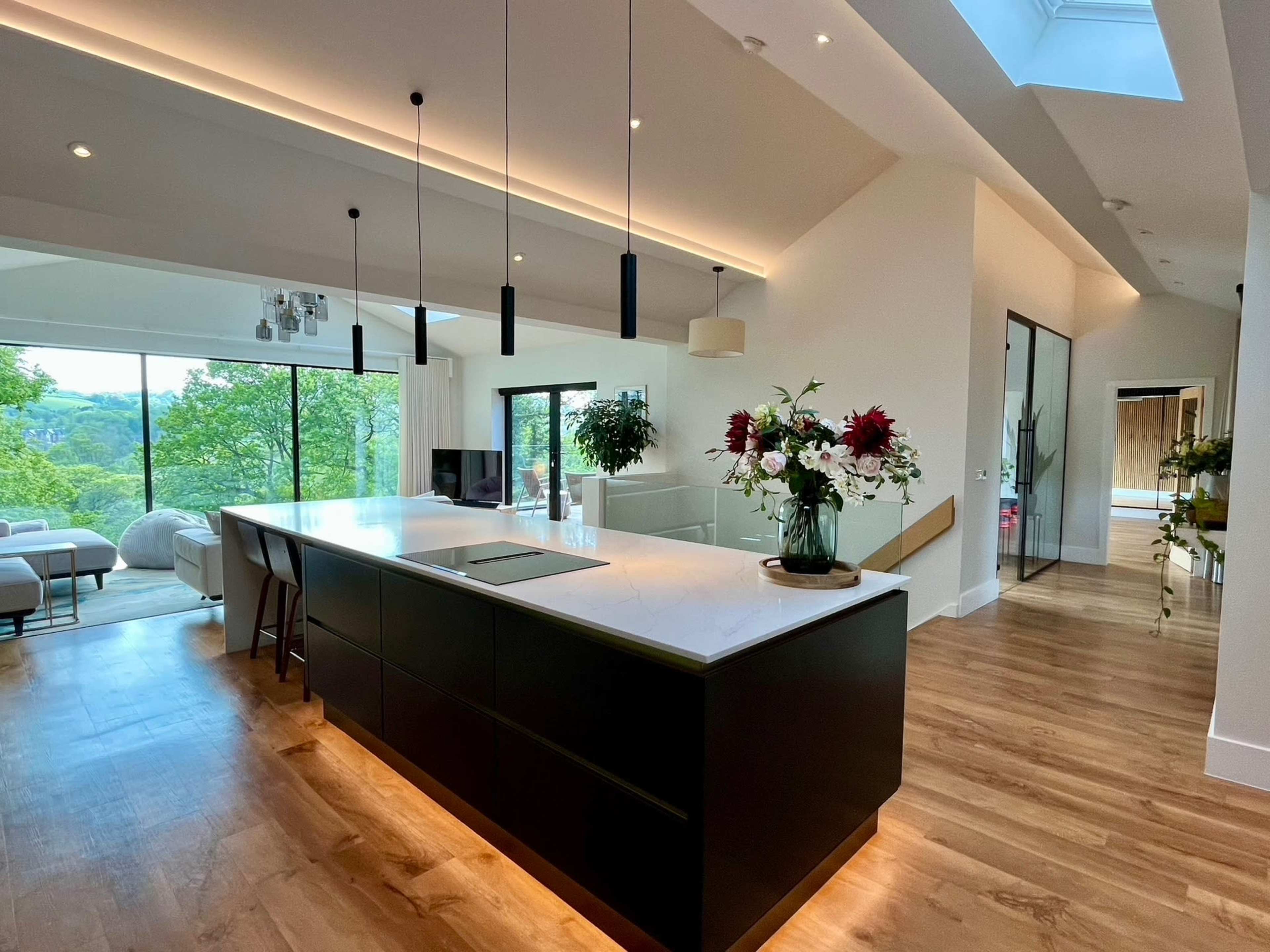 The image shows a modern kitchen with a large island, featuring a dark cabinetry and marble countertop, overlooking a spacious living area with large windows allowing natural light and views of greenery.