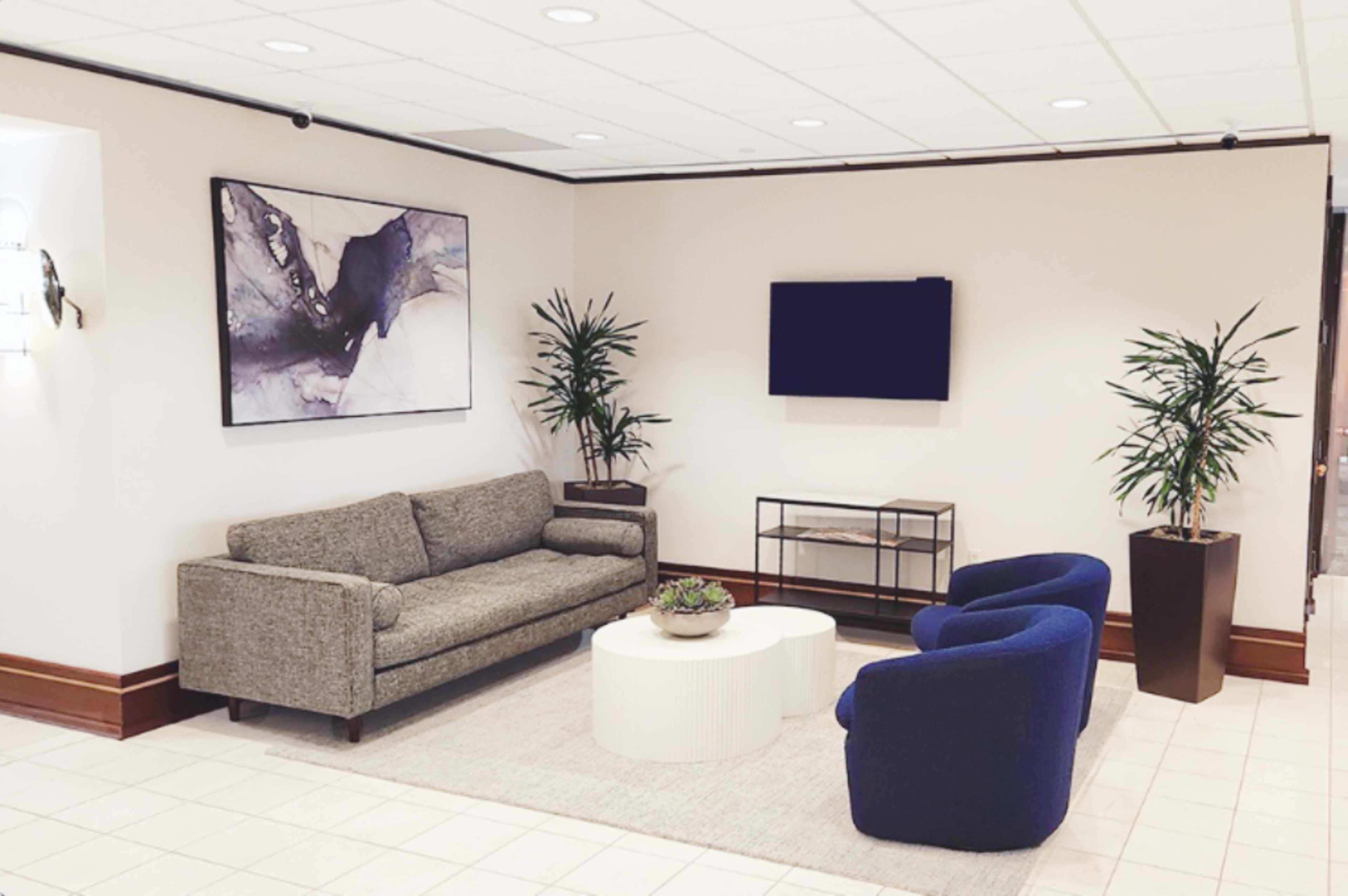 A modern waiting area features a gray sofa, two blue chairs, a circular coffee table, and potted plants, with abstract art and a television on the wall.
