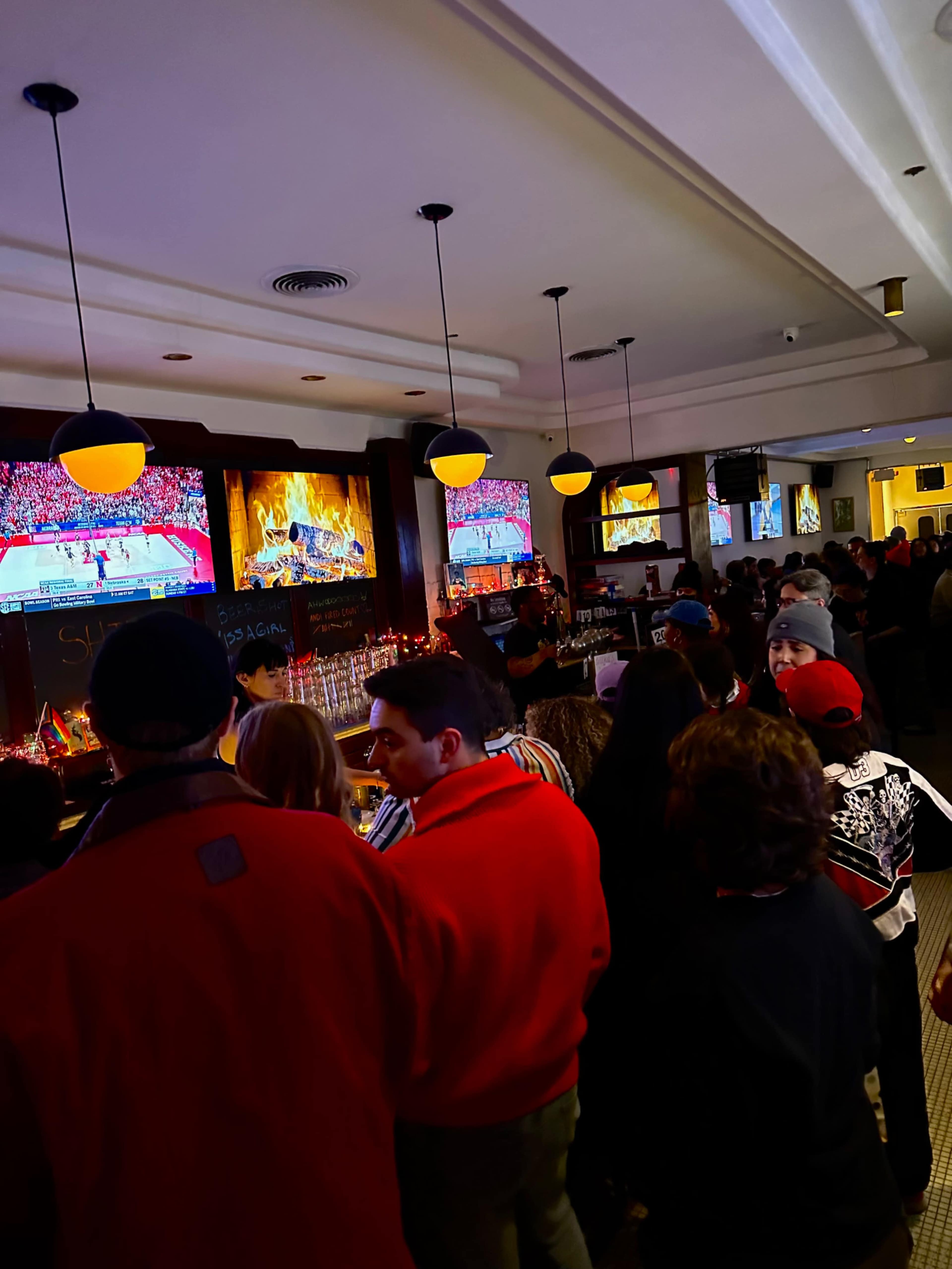 A crowded bar scene features patrons watching multiple TVs displaying a sports event, with a warm ambiance enhanced by decorative lights.