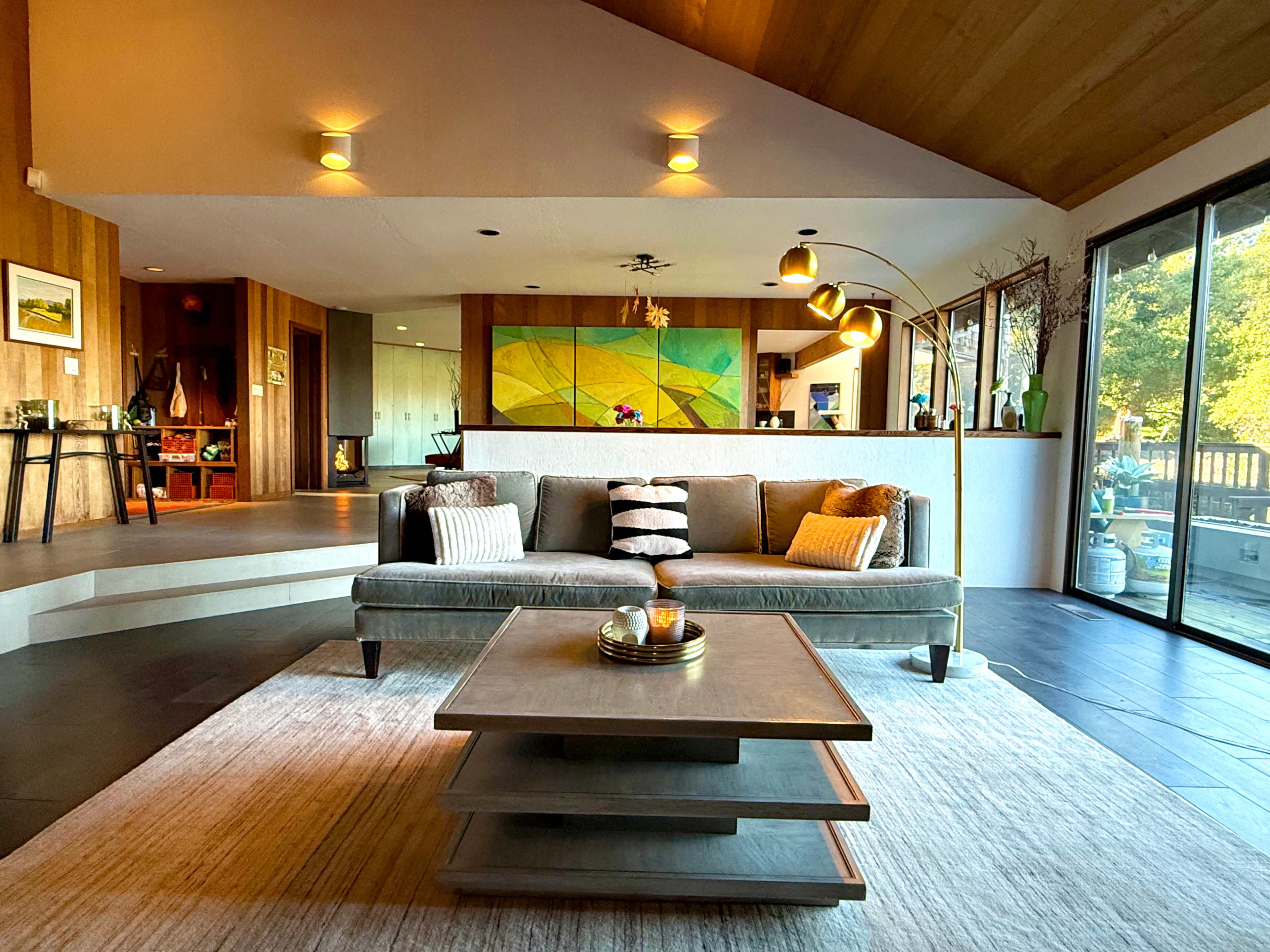 The image shows a modern living room with a gray sofa, a coffee table, and large windows providing natural light, all set in a space with wood and stone elements.