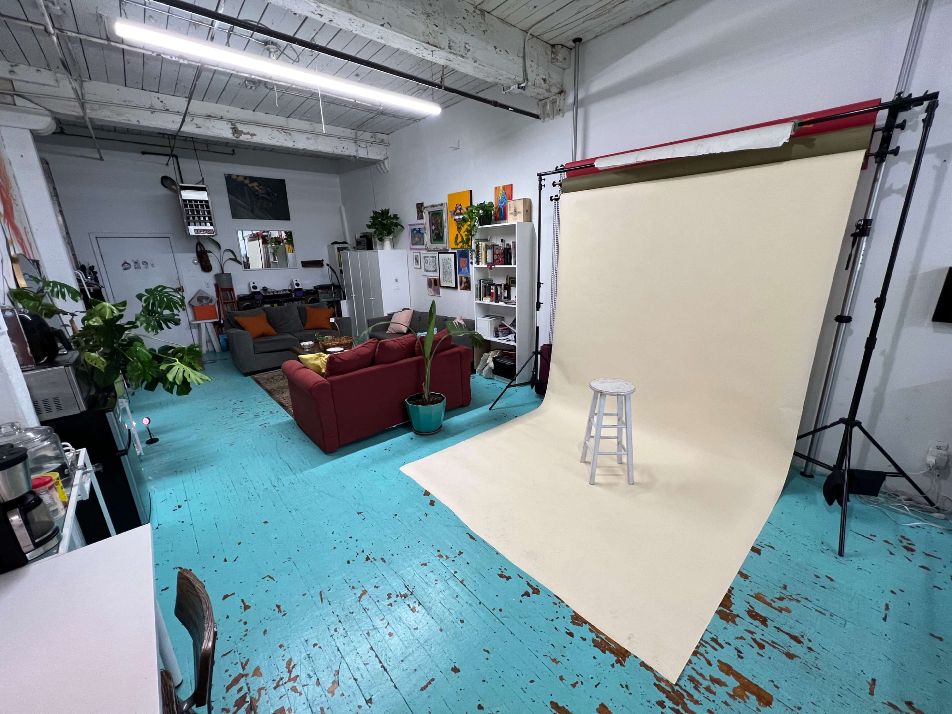 The image shows a spacious studio with a beige backdrop on a stand, a white stool in front, and a cozy seating area with a red sofa and various plants.