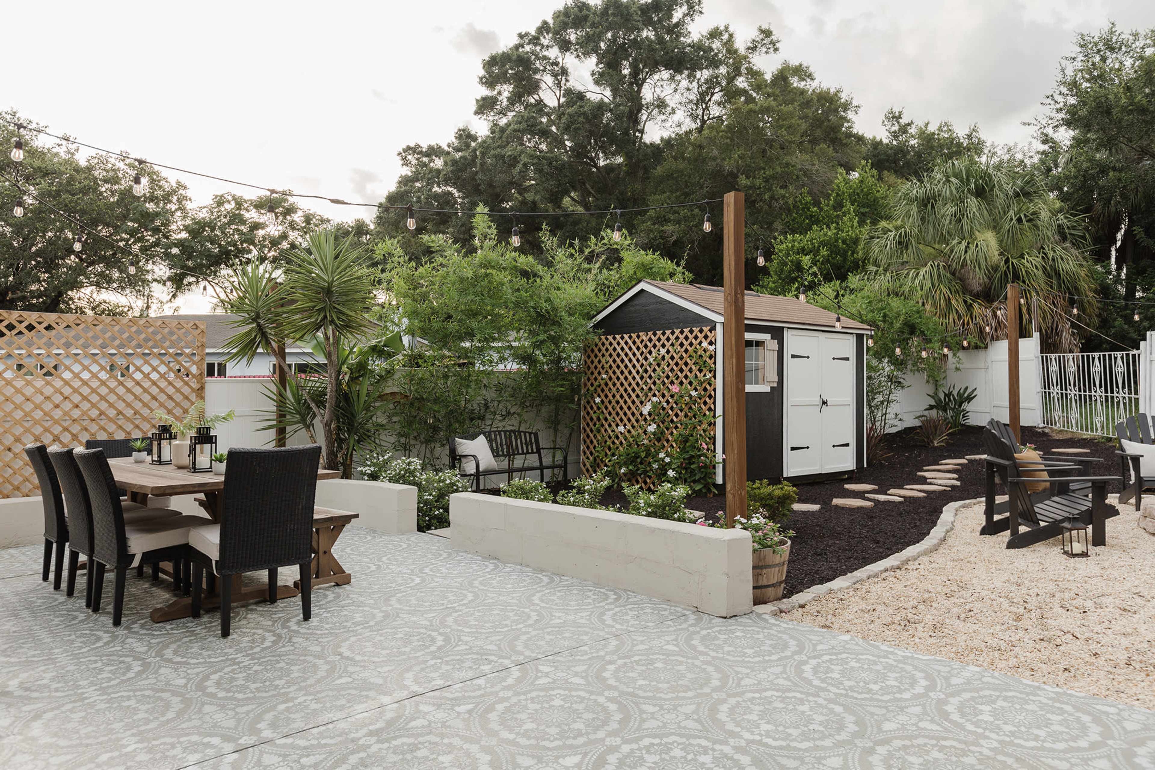 A well-designed backyard with a dining set, a shed, lush greenery, and a pathway lined with gravel and plants.