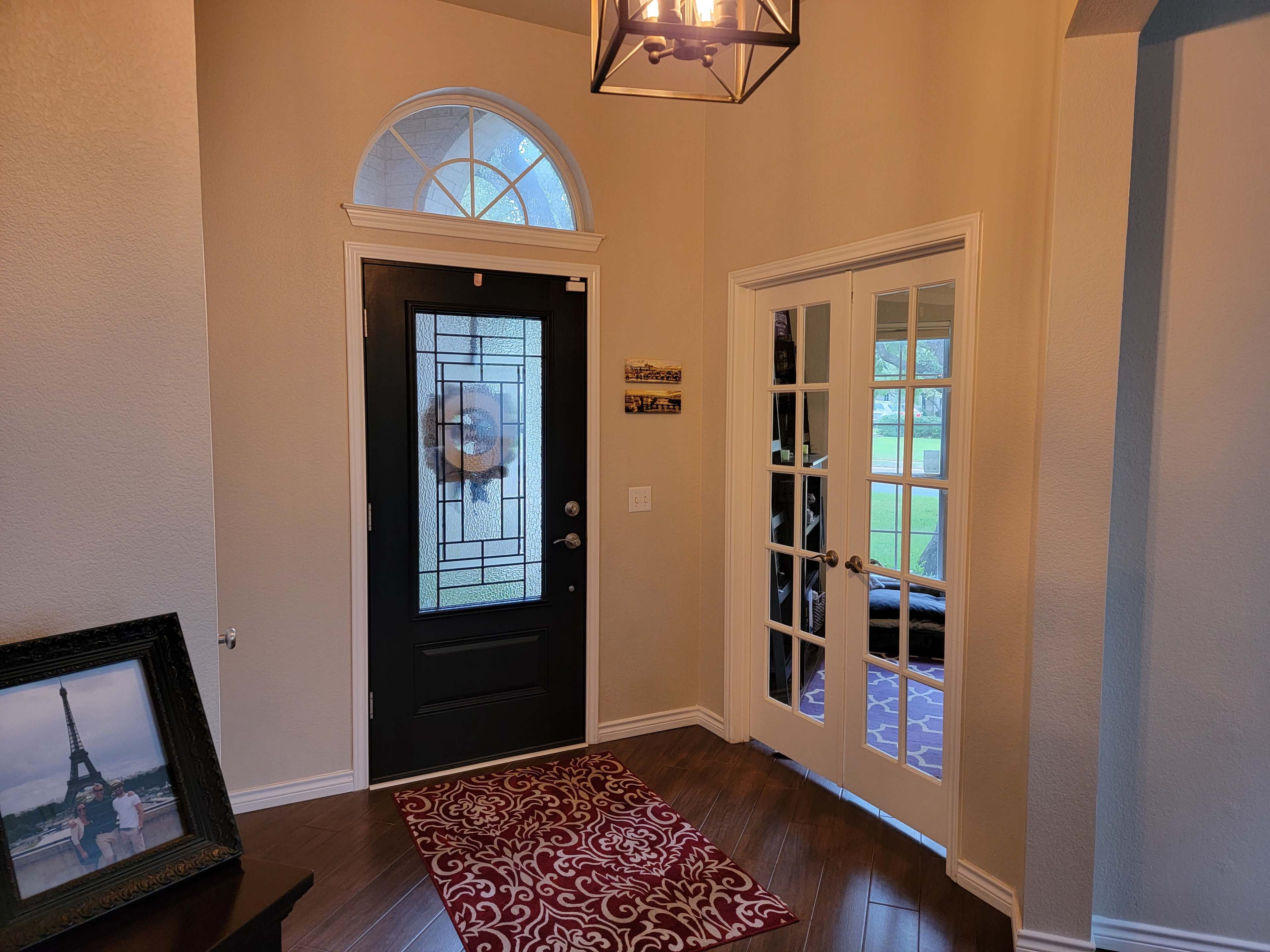 The image shows an entrance hall featuring a black front door with glass paneling, a chandelier, a decorative rug, and double French doors leading to an outdoor view.