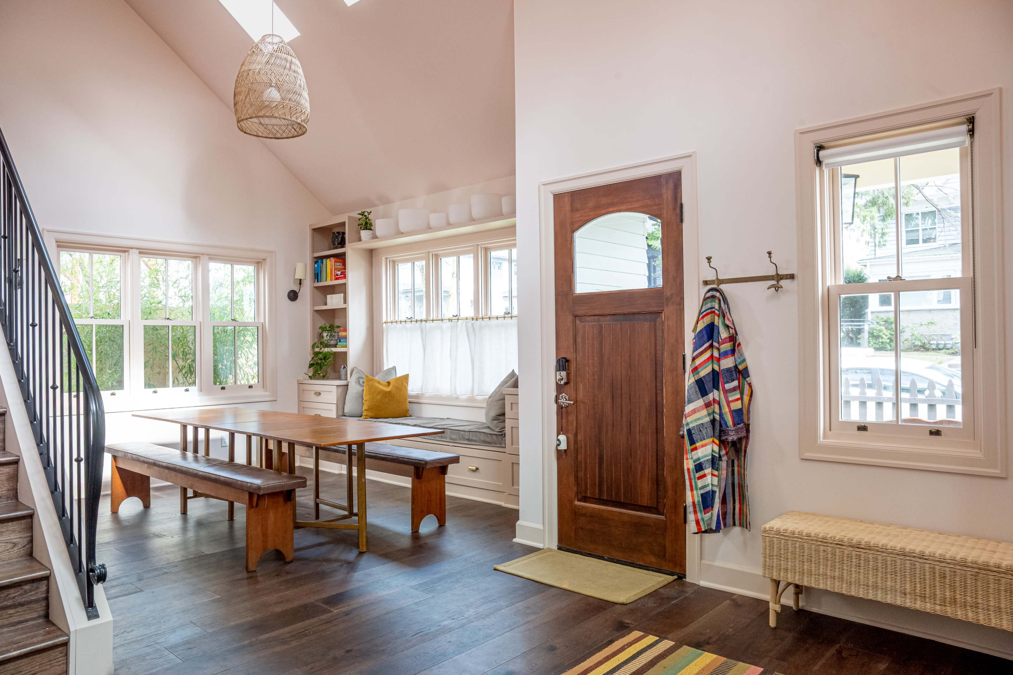 The interior of a home features a dining table and benches, a window seat with cushions, a wooden front door, and a staircase with a black railing.