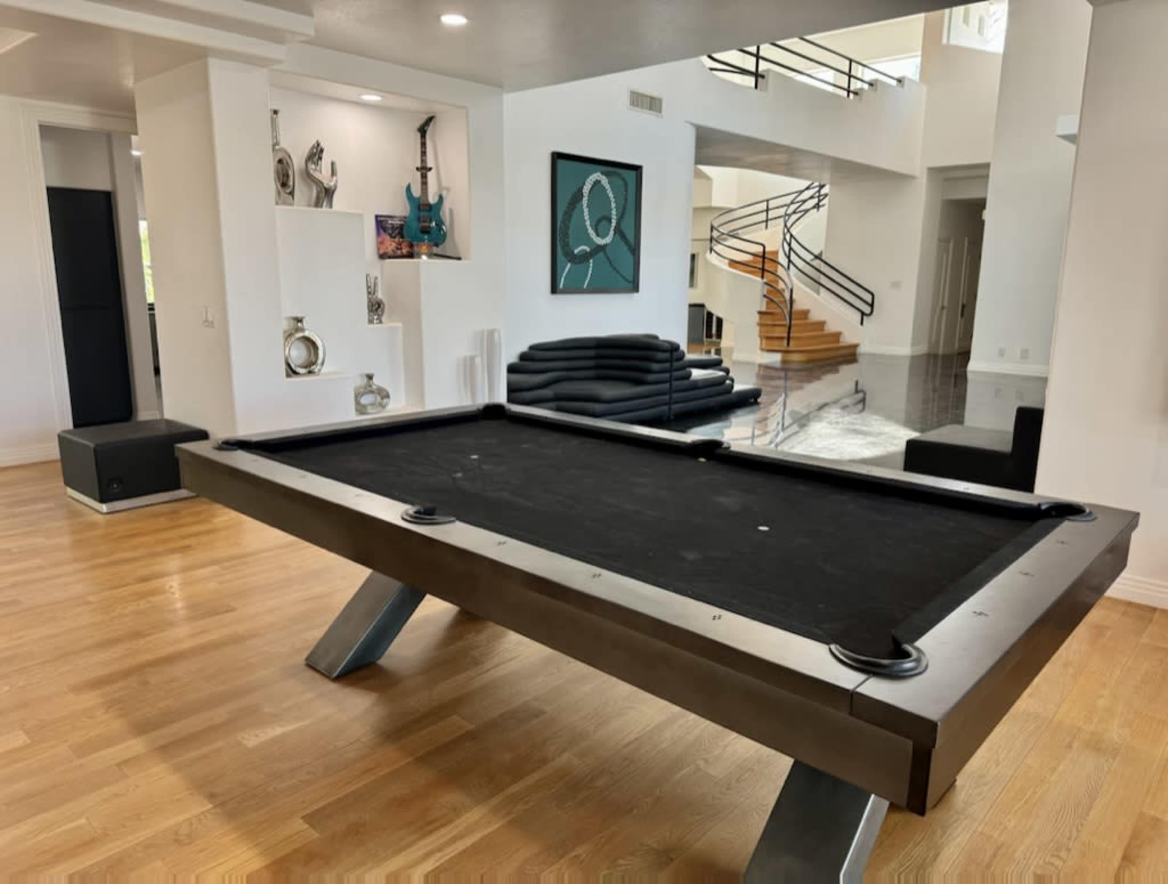 The image shows a modern interior with a sleek billiard table at the center, surrounded by minimalist furniture and a staircase leading to an upper level.
