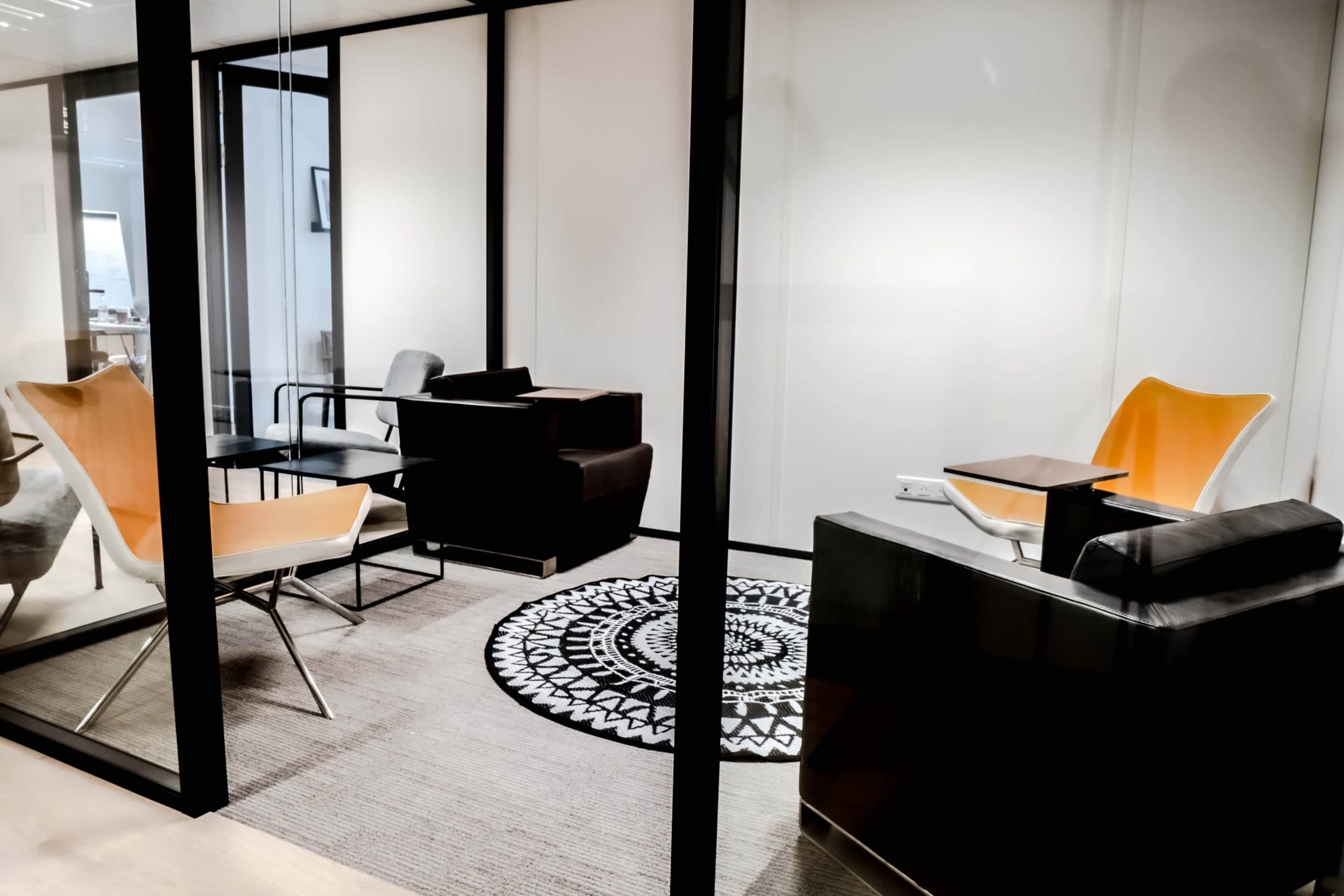 A modern office meeting space featuring orange-accented chairs, a circular black and white rug, and a contemporary desk within glass walls.