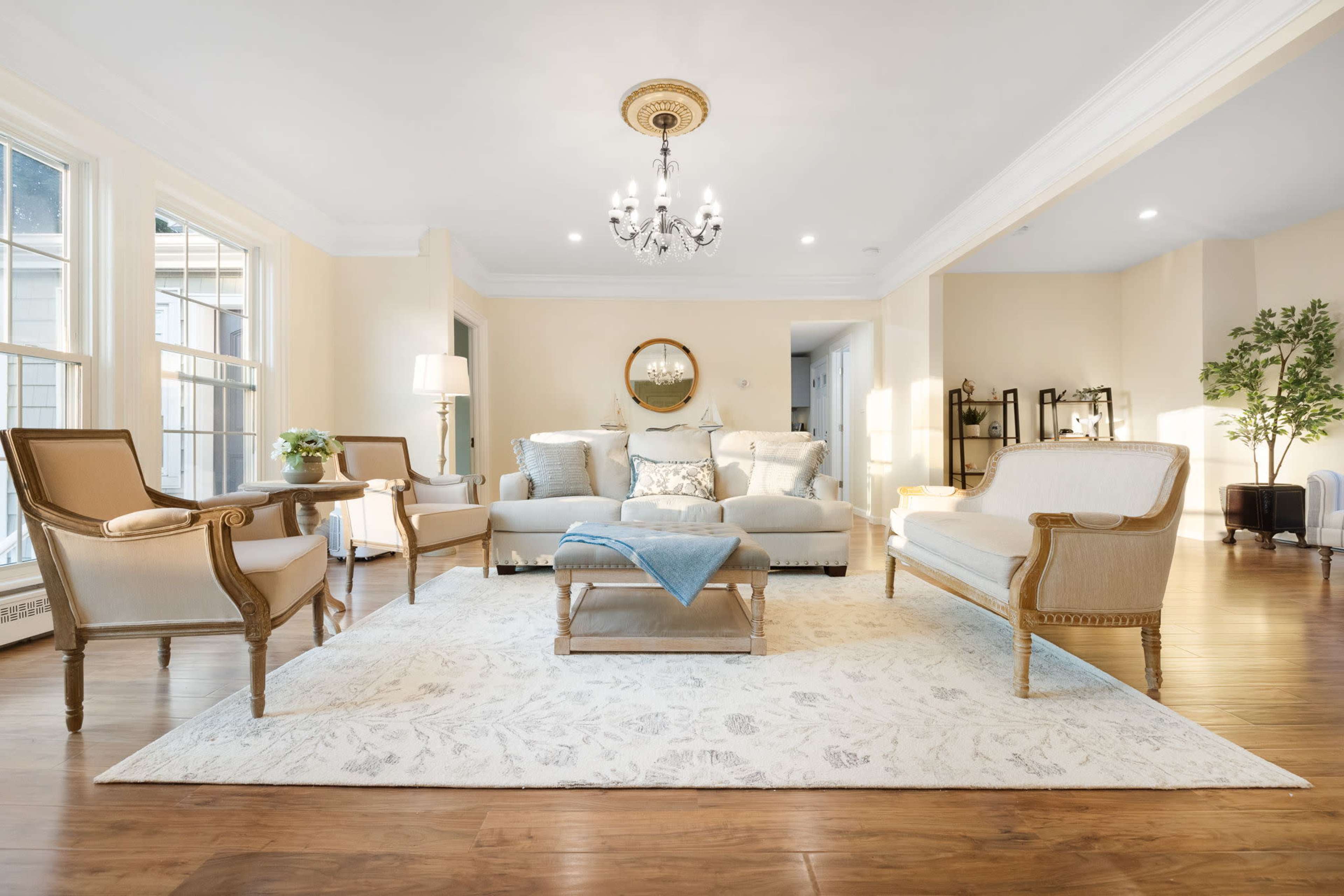 A well-lit living room with beige walls, hardwood flooring, and a central seating area comprising two armchairs, a sofa, and a coffee table on a decorative rug.