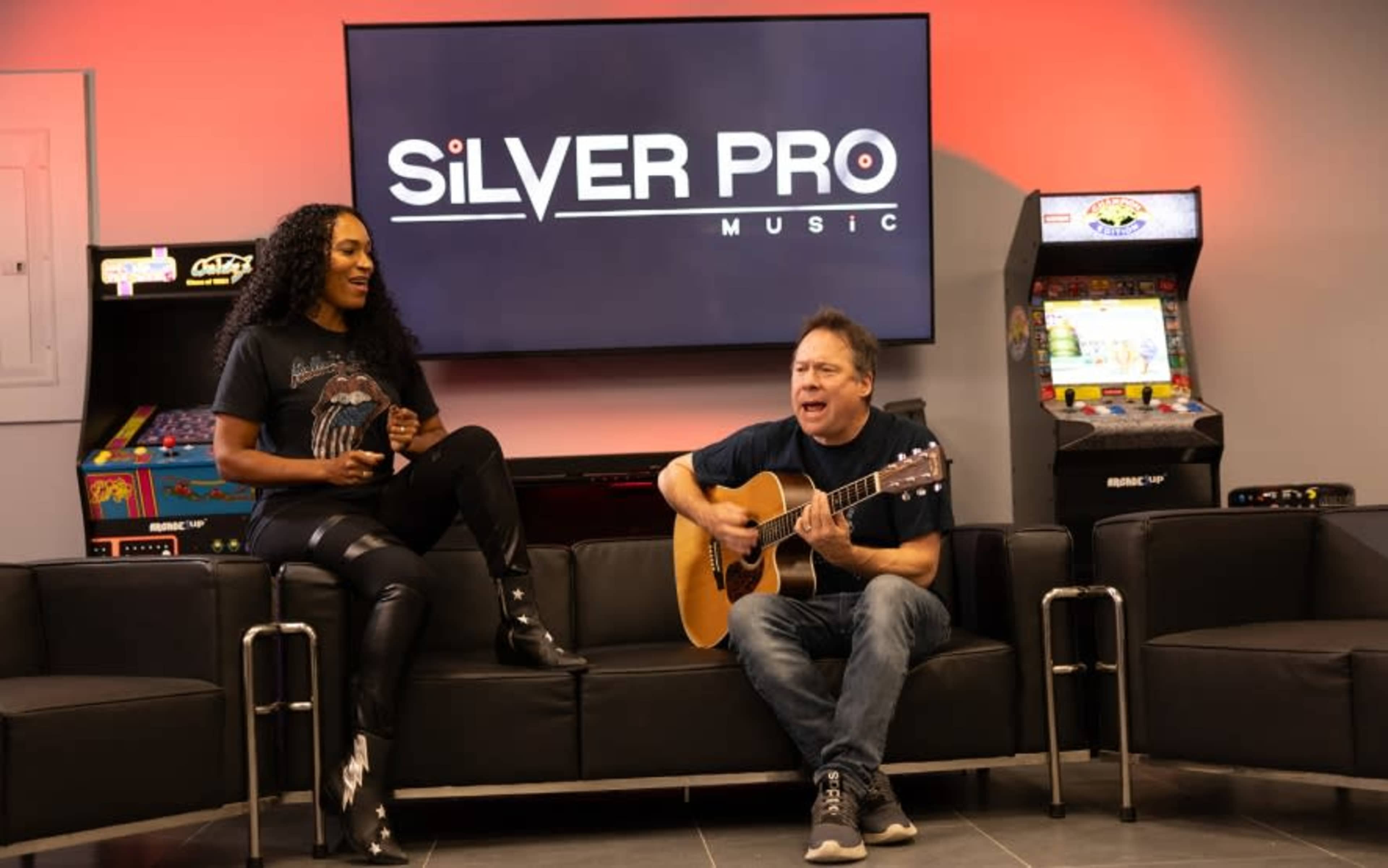 A woman performs vocally while a man plays guitar in a music studio adorned with arcade games and a large logo for Silver Pro Music in the background.
