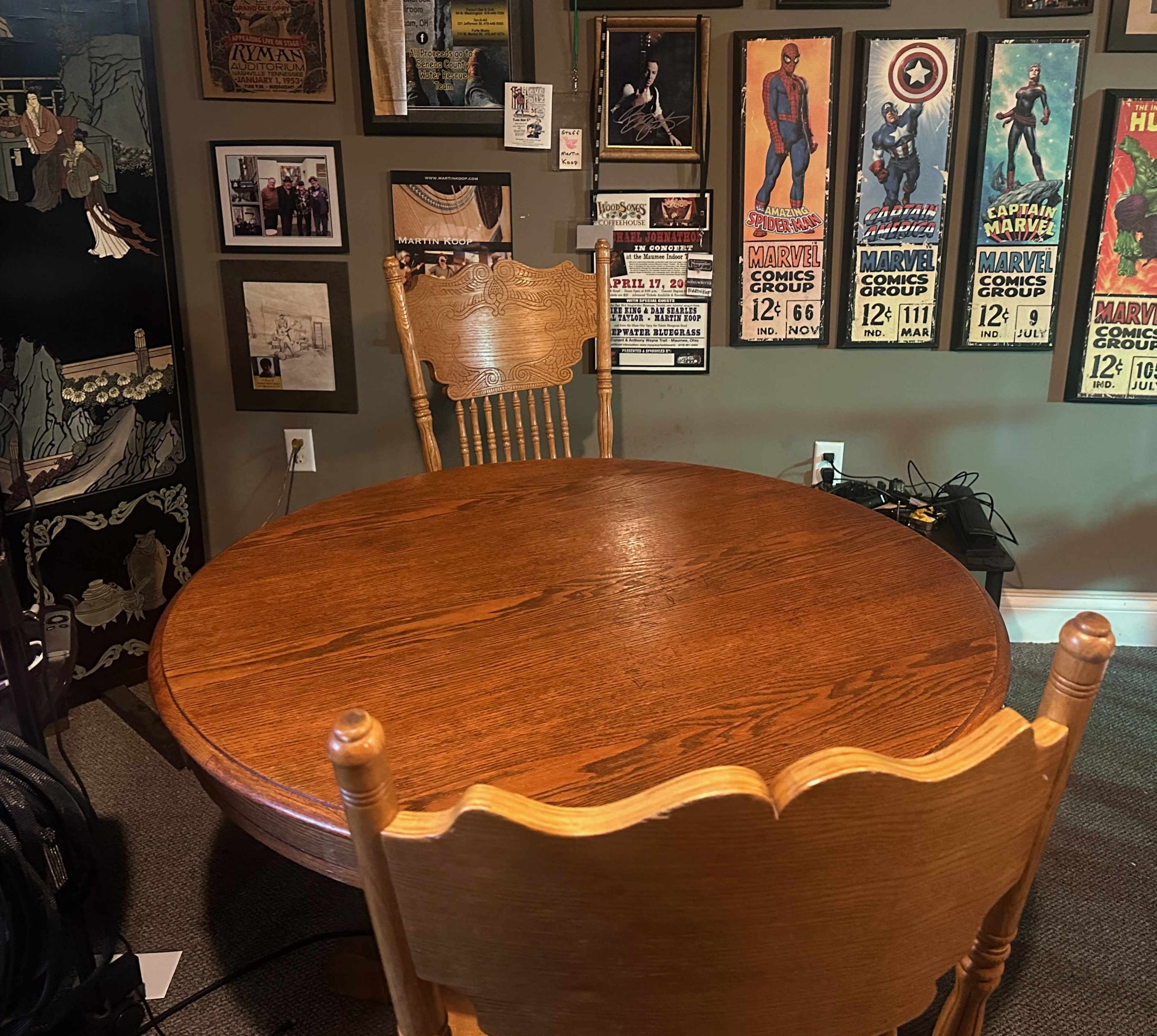 The image shows a wooden round dining table with one empty chair, surrounded by framed posters and photographs on the walls.