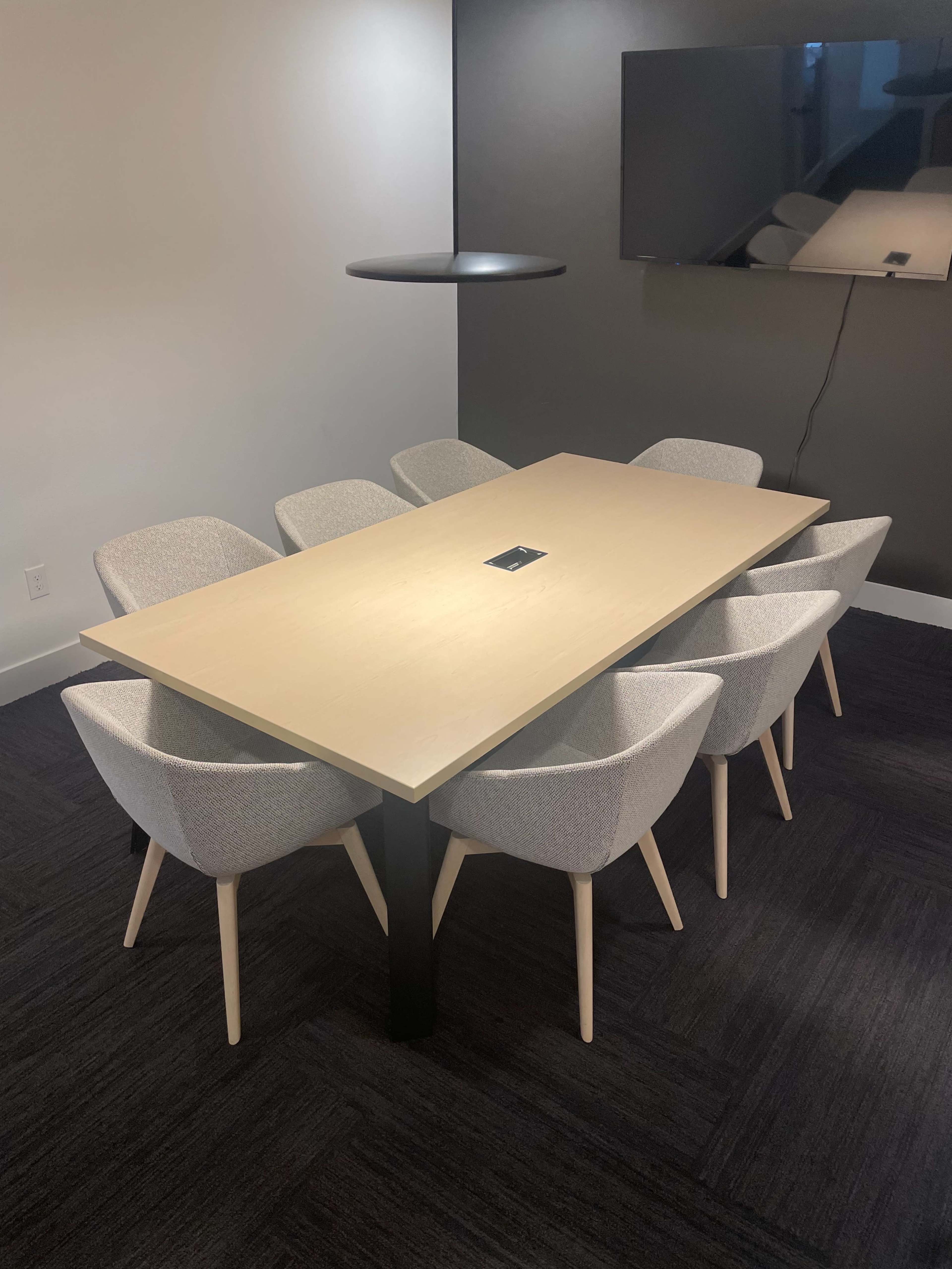 A modern conference room features a long wooden table surrounded by six upholstered chairs, with a large screen mounted on the wall.
